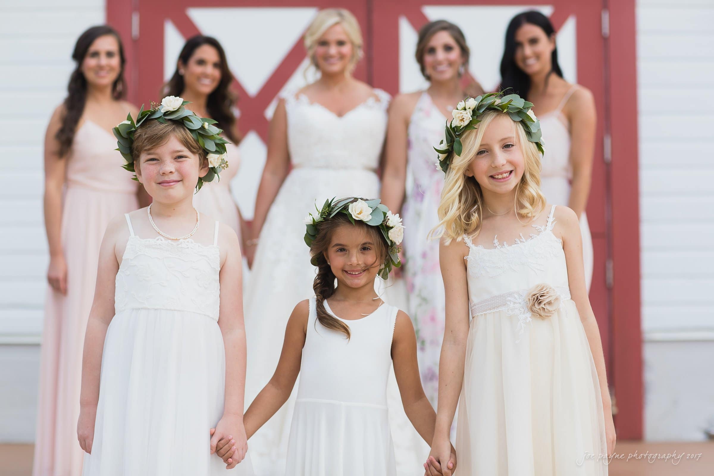 winmock nc wedding photographer - hallie & cullen Three young girls in white dresses with flower crowns stand holding hands in front of a group of six women wearing various pastel dresses. The background features a white wall with red barn doors.