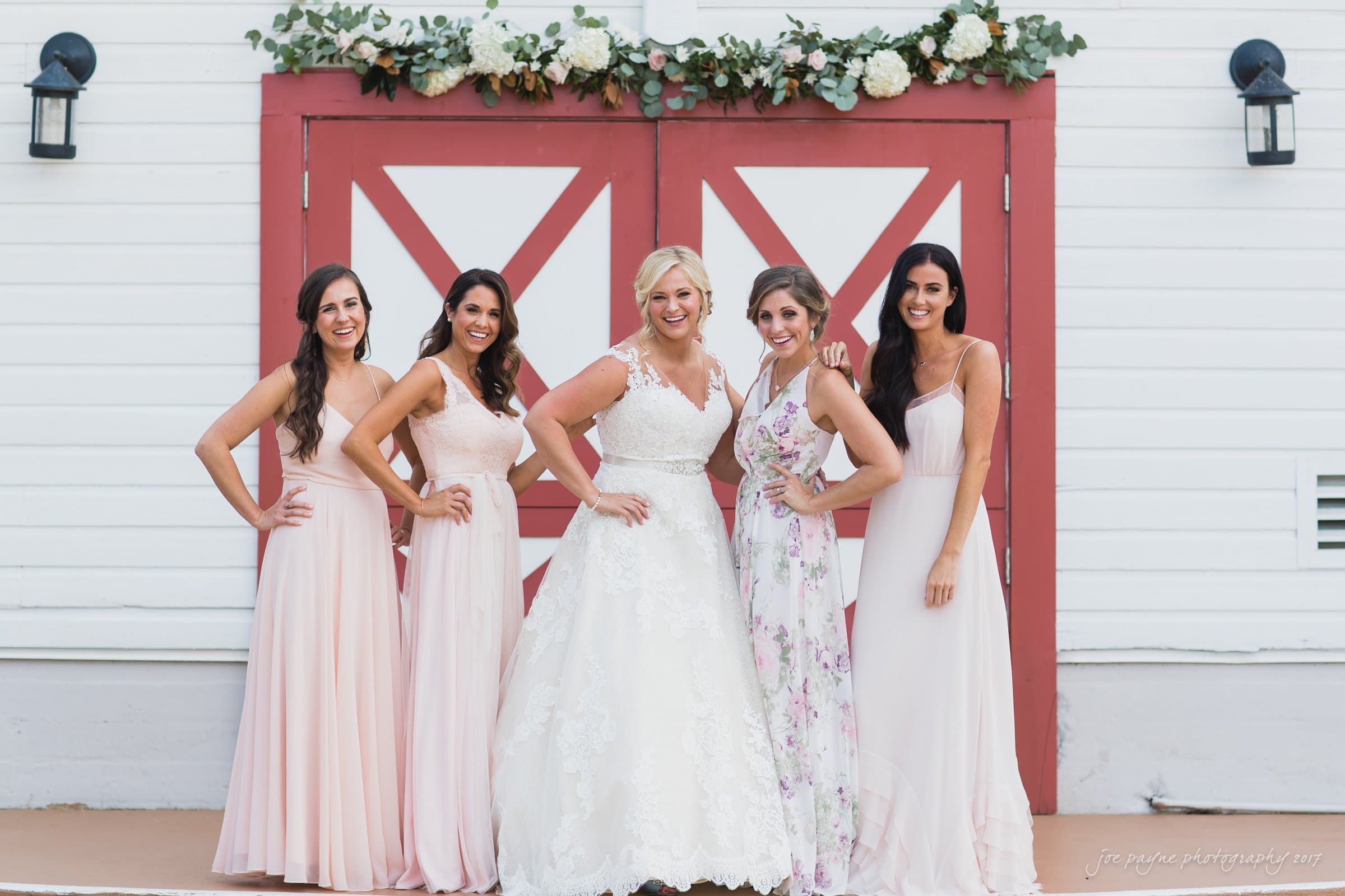 winmock nc wedding photographer - hallie & cullen A bride in a white gown stands with four bridesmaids in pastel dresses. They are posing in front of a red and white barn door adorned with floral decor. Each has a hand on their hip, smiling at the camera.