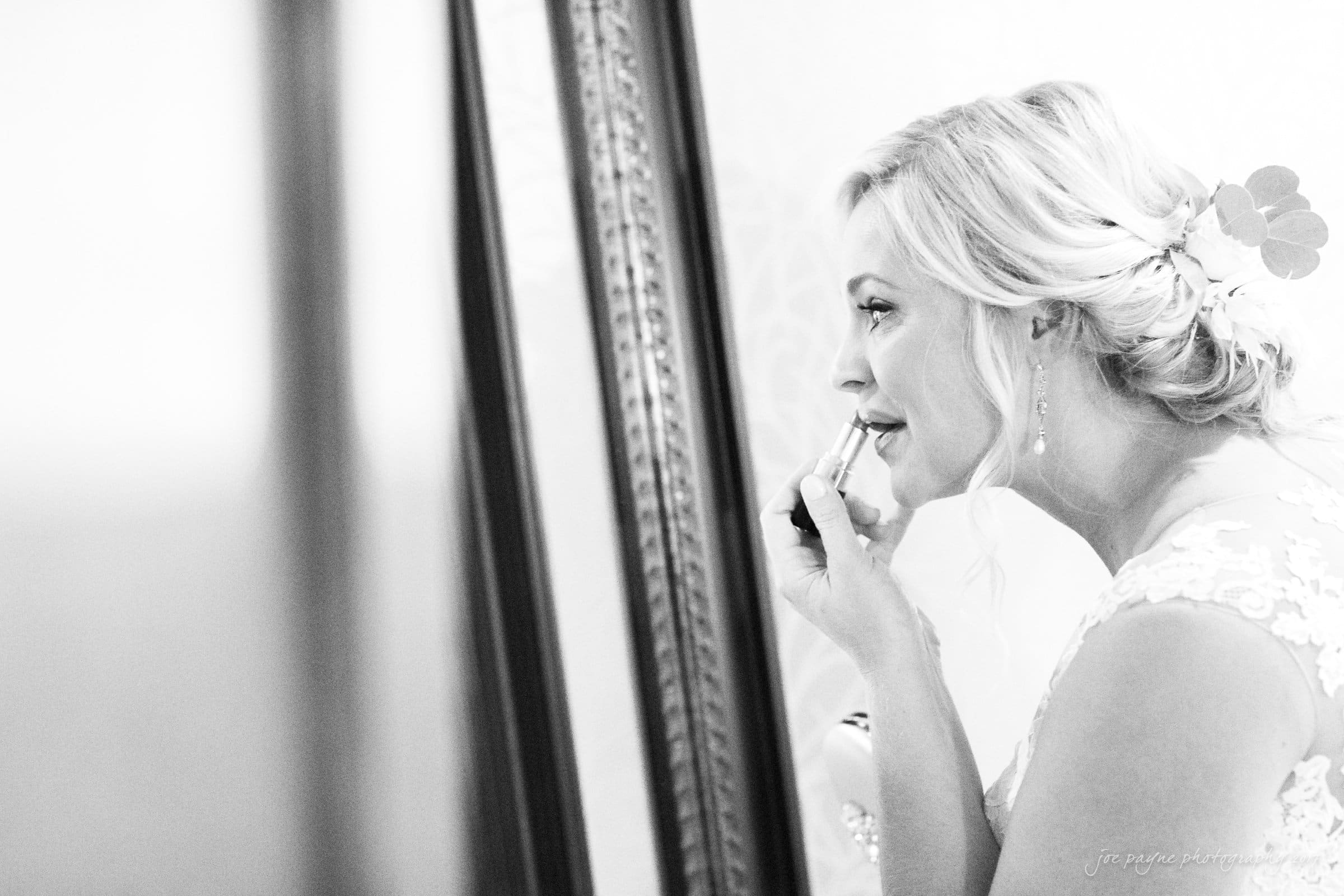 winmock nc wedding photographer - hallie & cullen A bride with blonde hair applies lipstick while looking in a mirror. She wears a lace dress and has a flower in her hair. The setting is elegant, and her expression is calm and focused. The photo is in black and white.