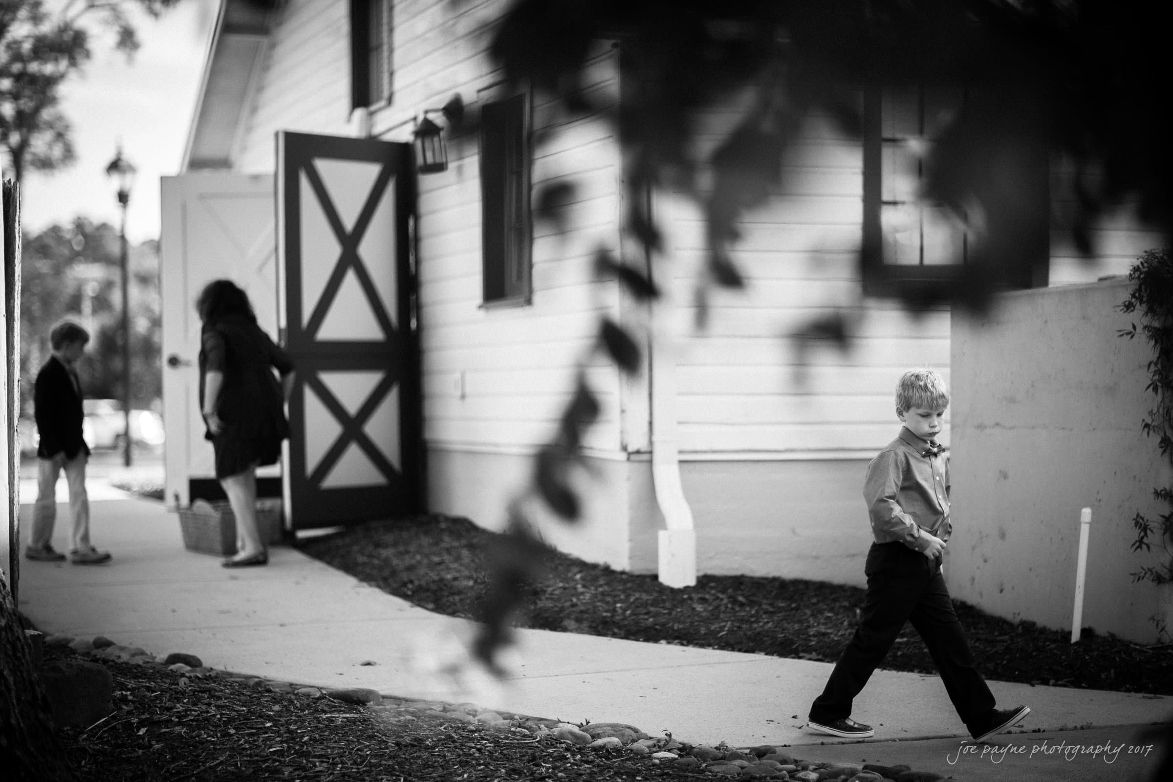 winmock nc wedding photographer - hallie & cullen A black and white photo of a young boy walking on a path near a white building with an open door. Two other people, a child and an adult, are near the entrance, facing away. Leaves are partially visible in the foreground.