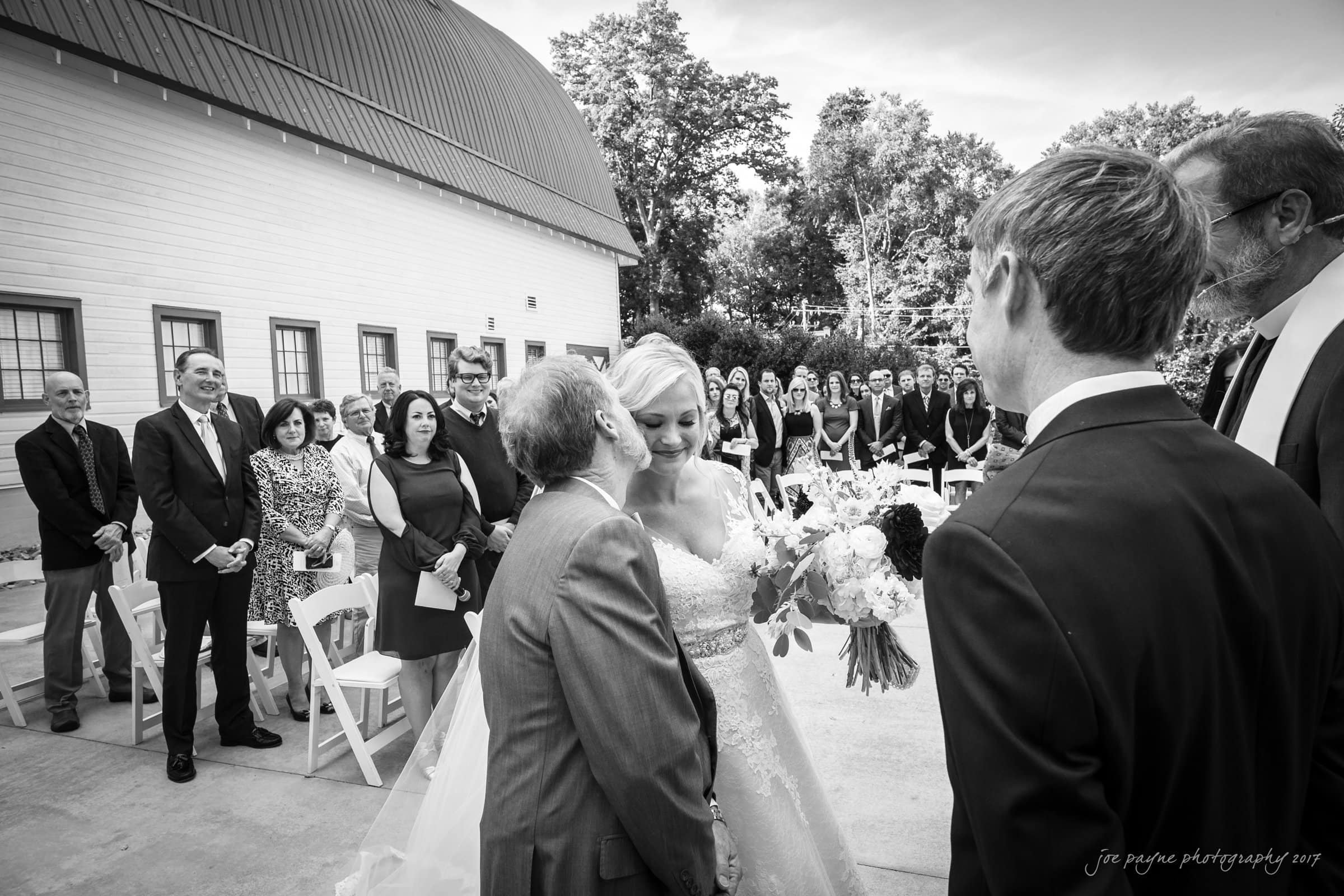 winmock nc wedding photographer - hallie & cullen A bride and her father share a heartfelt embrace during an outdoor wedding ceremony. Guests stand watching with smiles near a large, white barn. A groom stands facing the bride, holding a wedding bouquet.