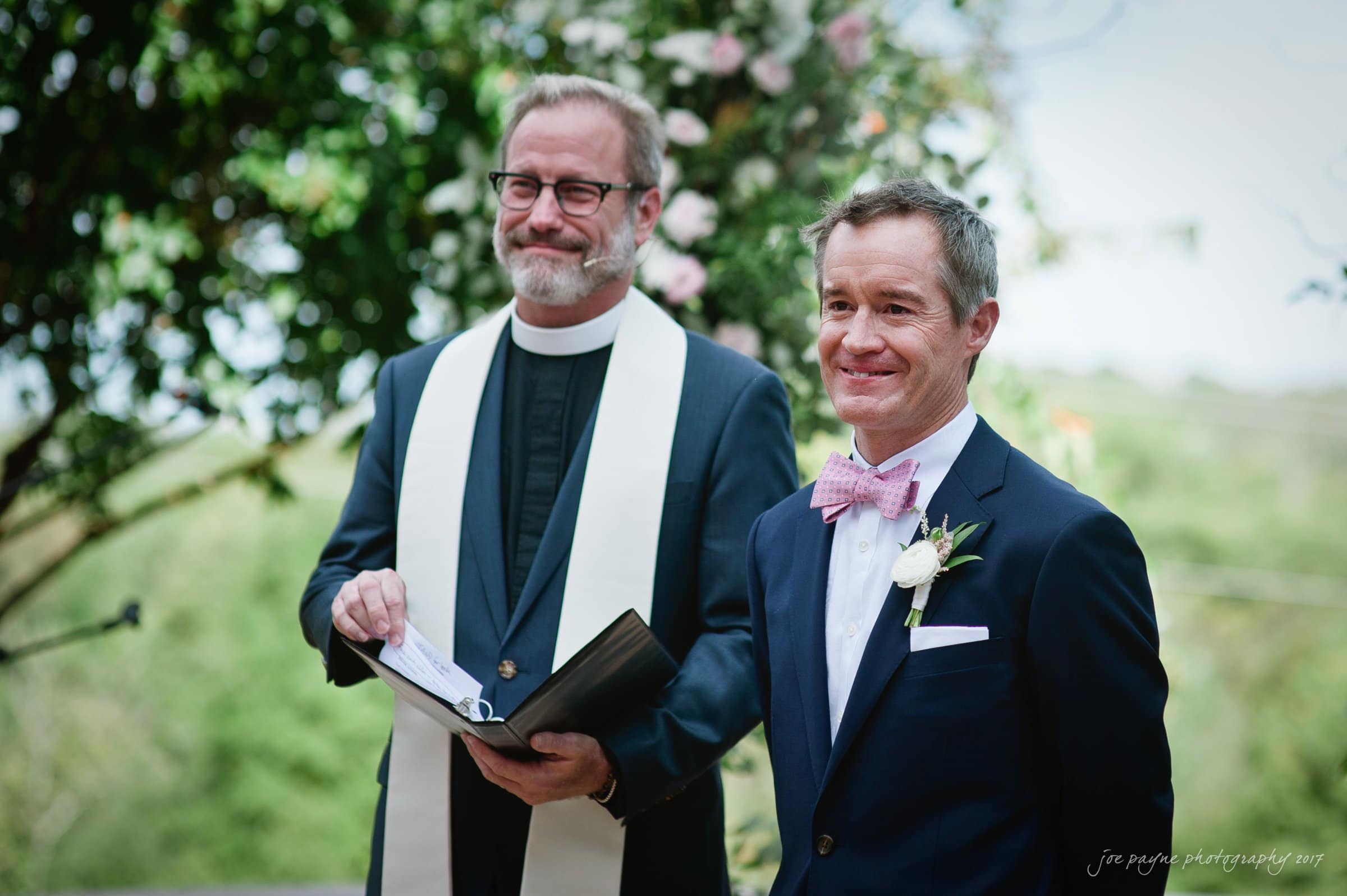 winmock nc wedding photographer - hallie & cullen A smiling officiant in a black robe and white stole stands beside a man in a dark suit with a pink bow tie, holding a book in an outdoor setting with greenery and flowers in the background.