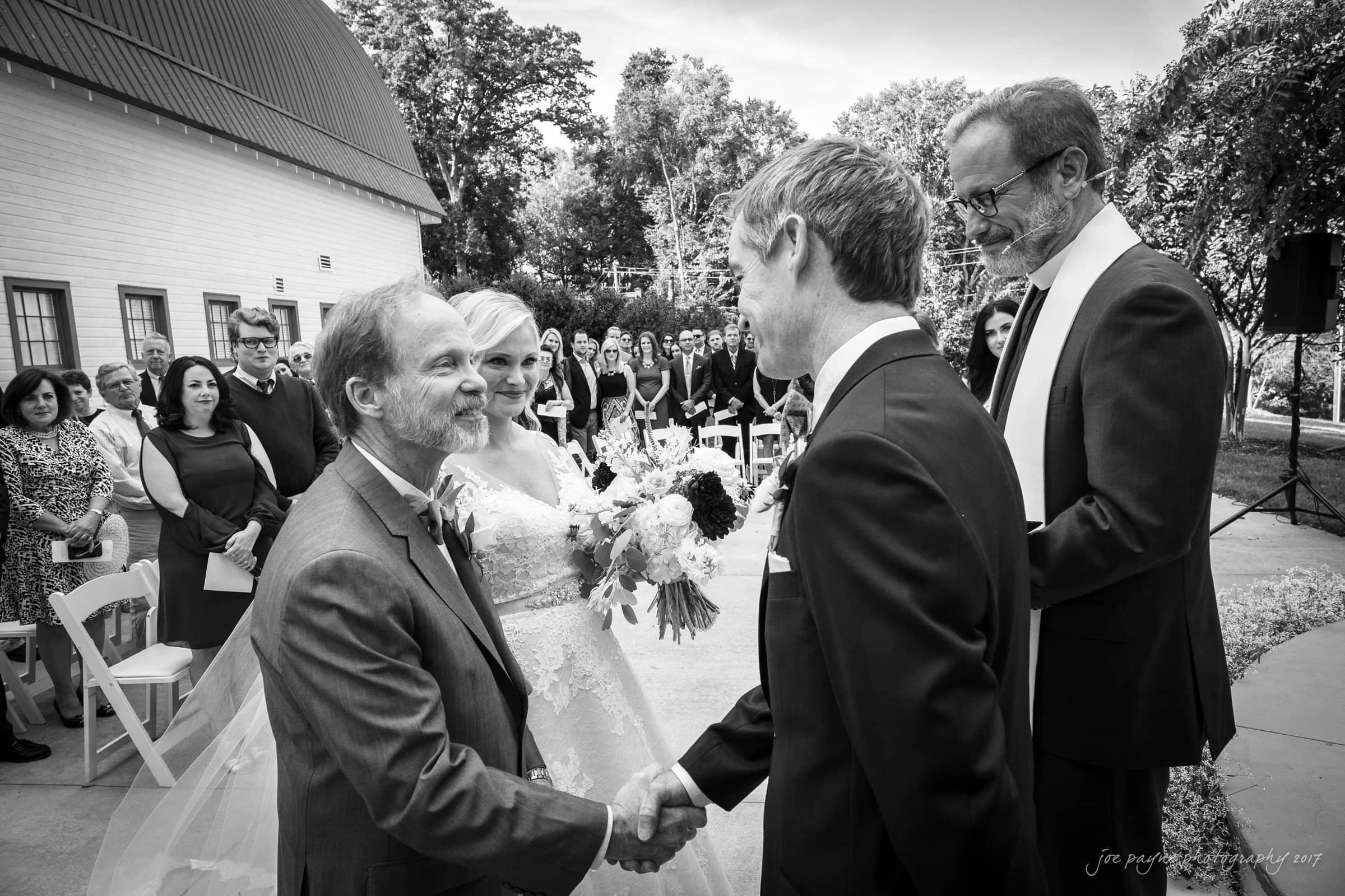 winmock nc wedding photographer - hallie & cullen A groom shakes hands with the brides father as the bride stands beside them, holding a bouquet. The ceremony is outdoors with guests seated in the background. A man in a suit watches the exchange. Black and white photo.
