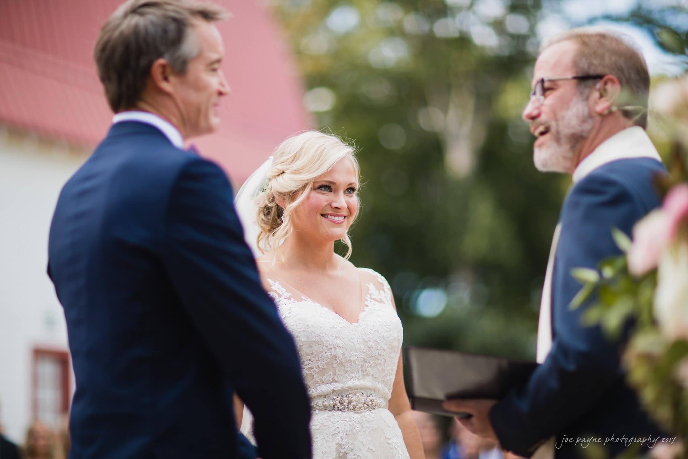 winmock nc wedding photographer - hallie & cullen A bride and groom stand facing each other during an outdoor wedding ceremony. The bride smiles as a bearded man officiates. The groom wears a dark suit. They are surrounded by greenery and an out-of-focus building in the background.