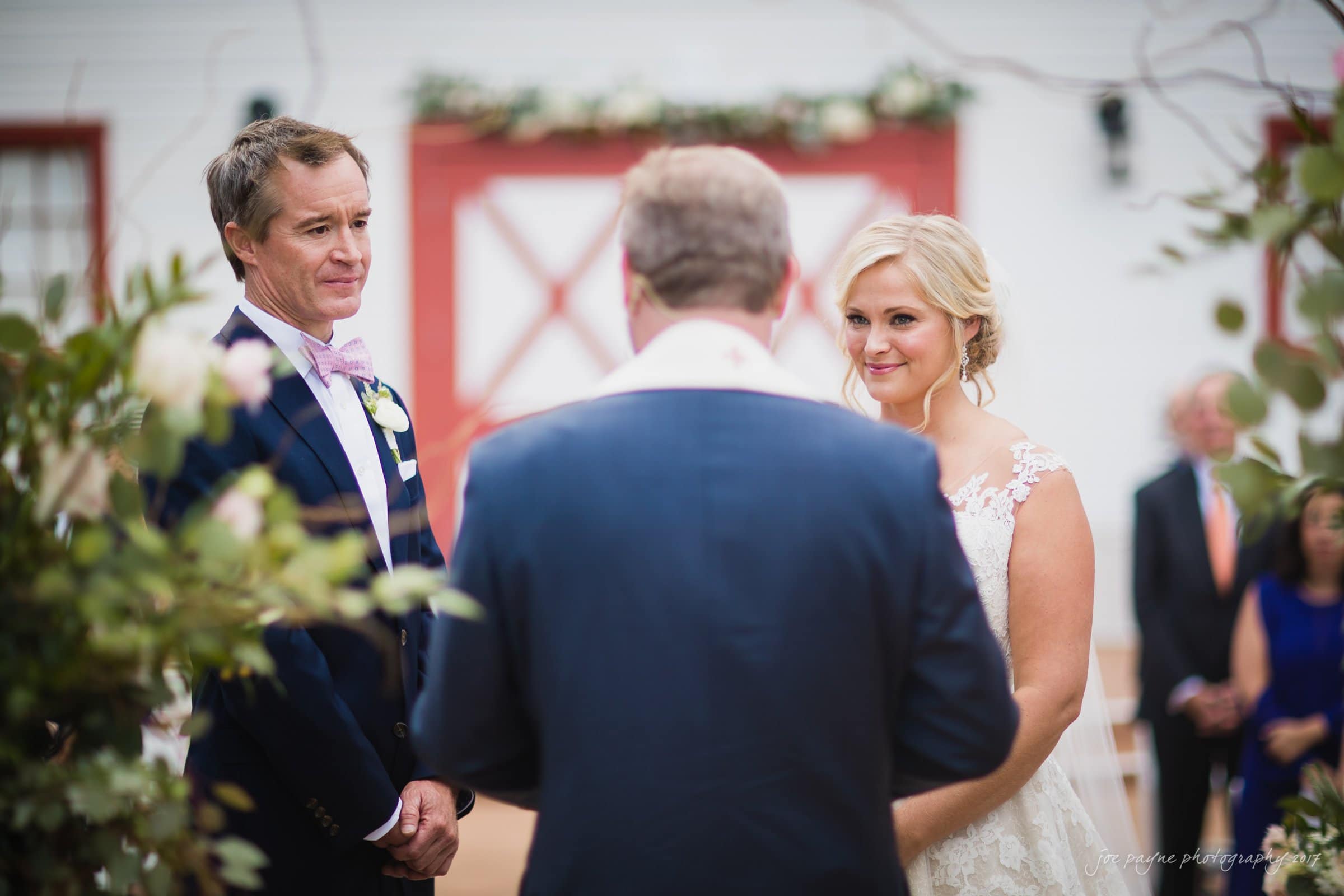 winmock nc wedding photographer - hallie & cullen A bride and groom stand facing an officiant during their outdoor wedding ceremony. The groom wears a dark suit and the bride is in a white gown. The background shows a building with red doors and floral decorations.