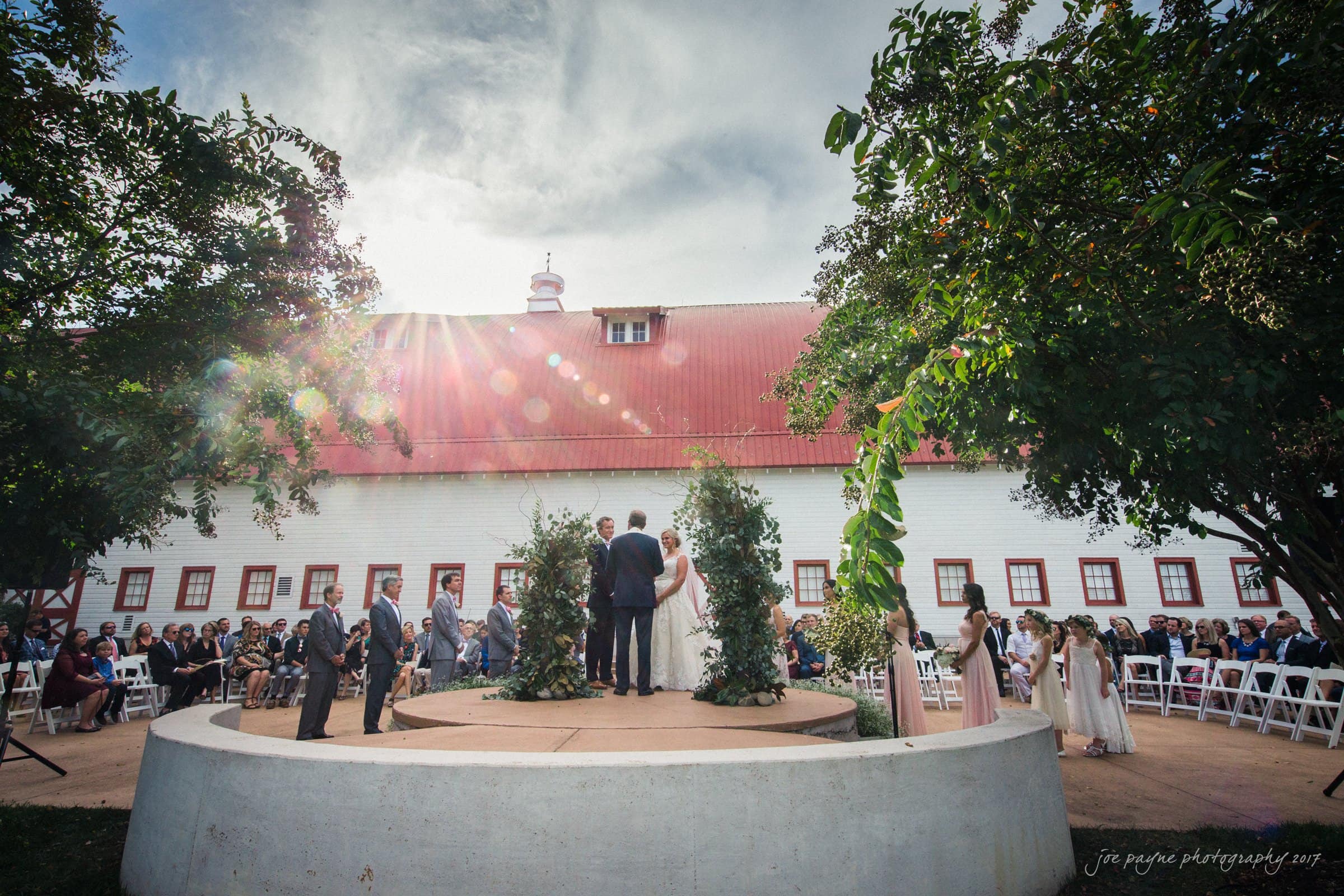 winmock nc wedding photographer - hallie & cullen A couple stands at an outdoor wedding ceremony in front of a white barn with a red roof. Guests are seated on white chairs, and the scene is framed by trees. The sun shines brightly, creating a lens flare effect in the sky.