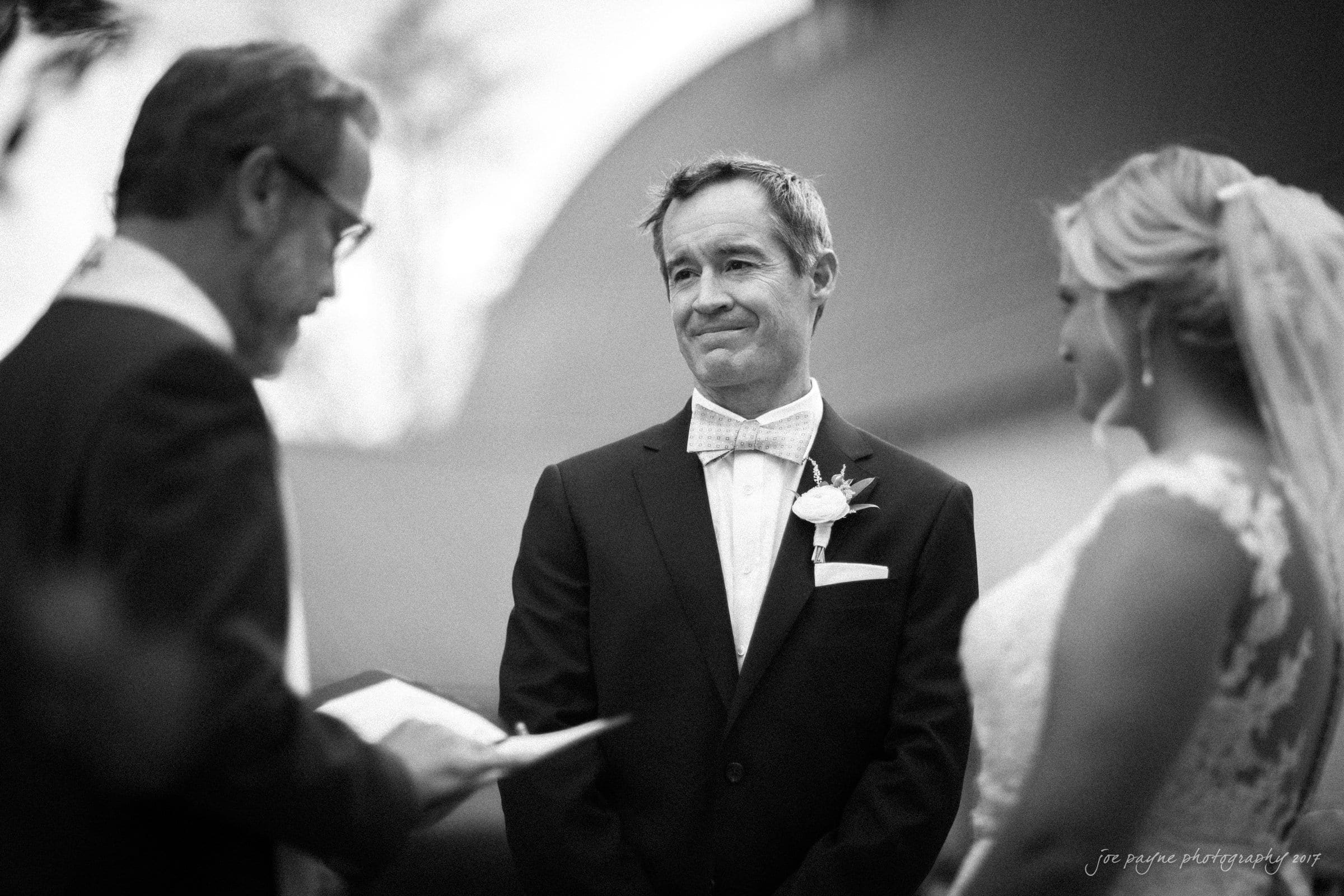 winmock nc wedding photographer - hallie & cullen A black and white photo of a wedding ceremony. A groom in a suit and bow tie stands smiling, looking at the bride in a lace gown. They are facing an officiant who is reading from a book. The mood is joyful and intimate.
