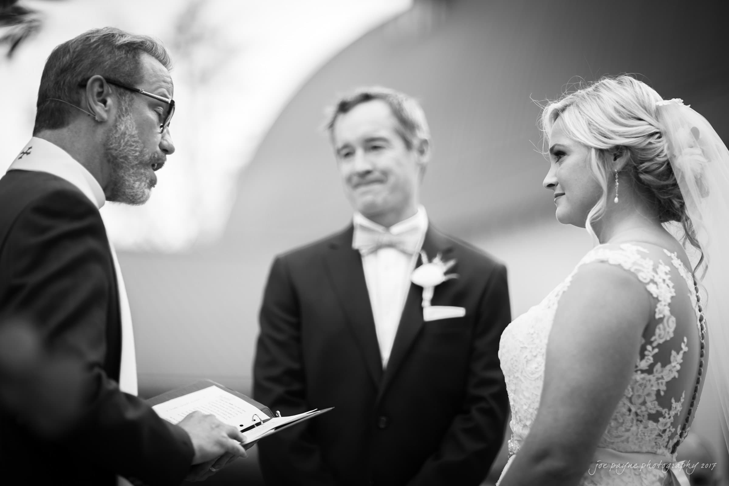 winmock nc wedding photographer - hallie & cullen A black and white photo of a wedding ceremony. A couple stands facing each other, smiling. The bride is in a lace dress and veil, and the groom wears a suit with a bow tie. An officiant holding a book conducts the ceremony.