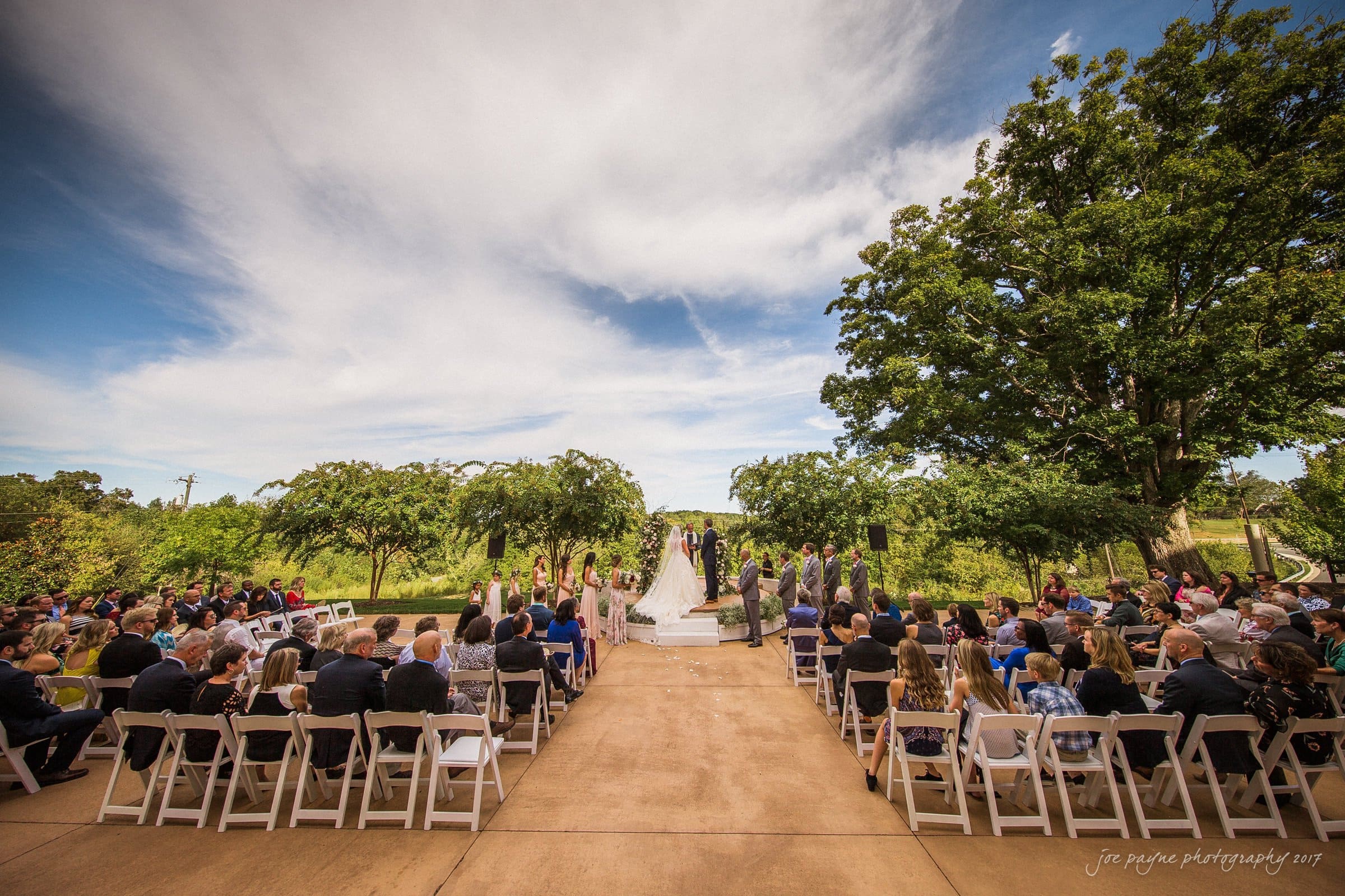 winmock nc wedding photographer - hallie & cullen A wedding ceremony takes place outdoors under a partly cloudy sky. Guests are seated on both sides of the aisle, lined with white chairs. The bride and groom stand at the altar, surrounded by greenery and trees.