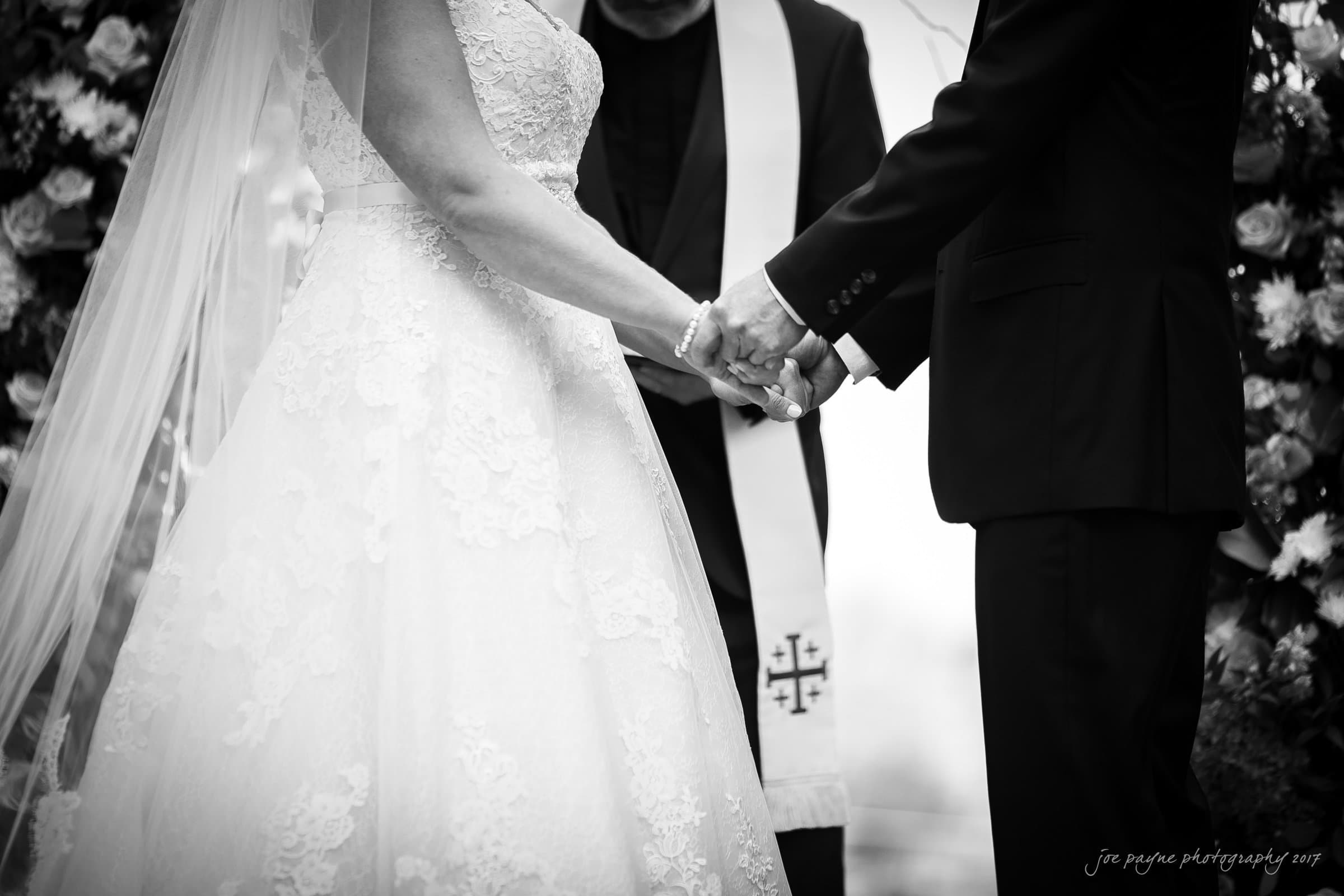 winmock nc wedding photographer - hallie & cullen A black and white image of a couple holding hands during a wedding ceremony. The bride is in a lace gown with a veil, and the groom is in a dark suit. A priest, wearing a stole with cross patterns, stands behind them.