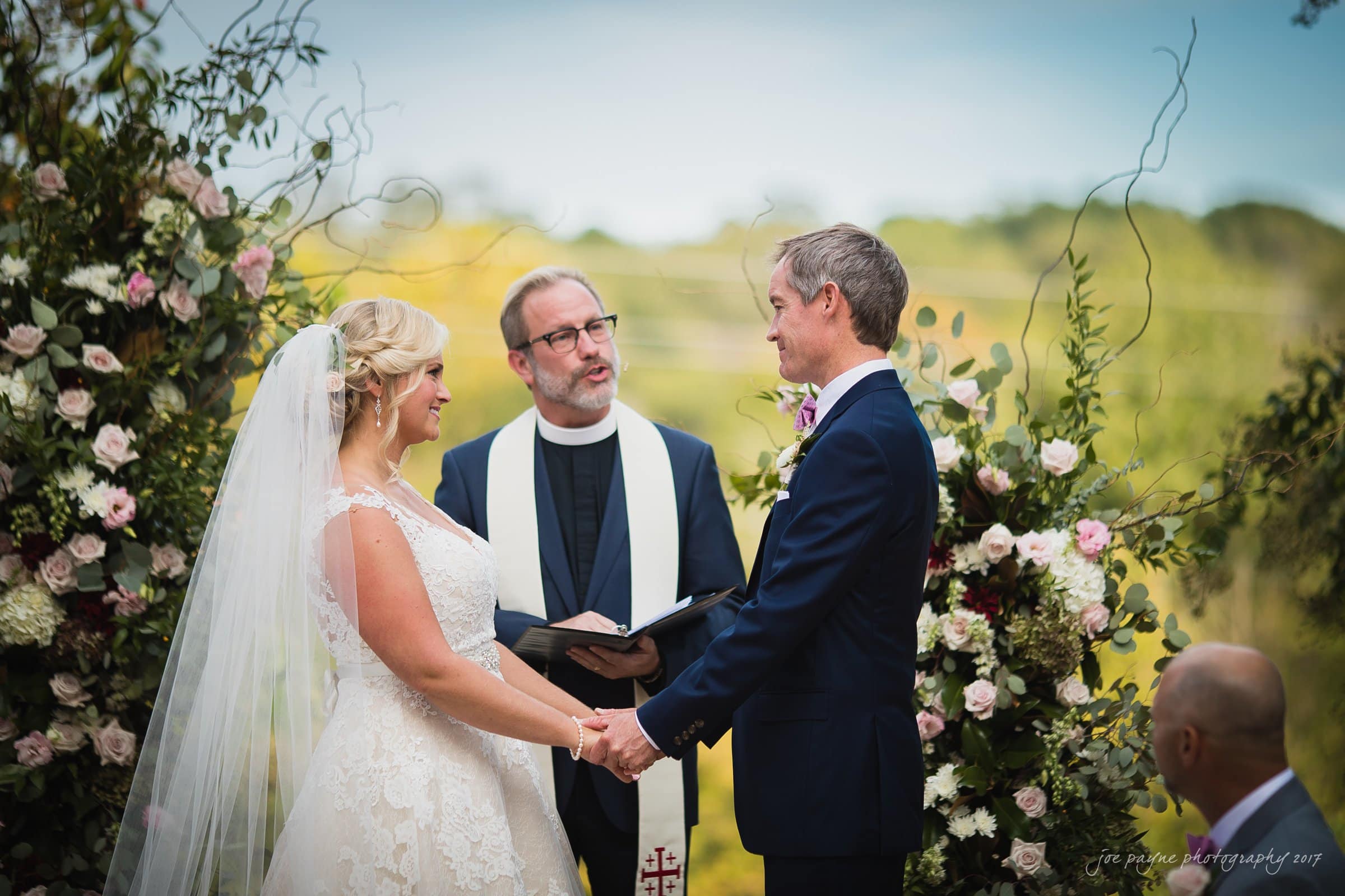 winmock nc wedding photographer - hallie & cullen A bride and groom stand holding hands facing each other during an outdoor wedding ceremony. A priest in the middle holds a book. The scene is adorned with beautiful floral arrangements.