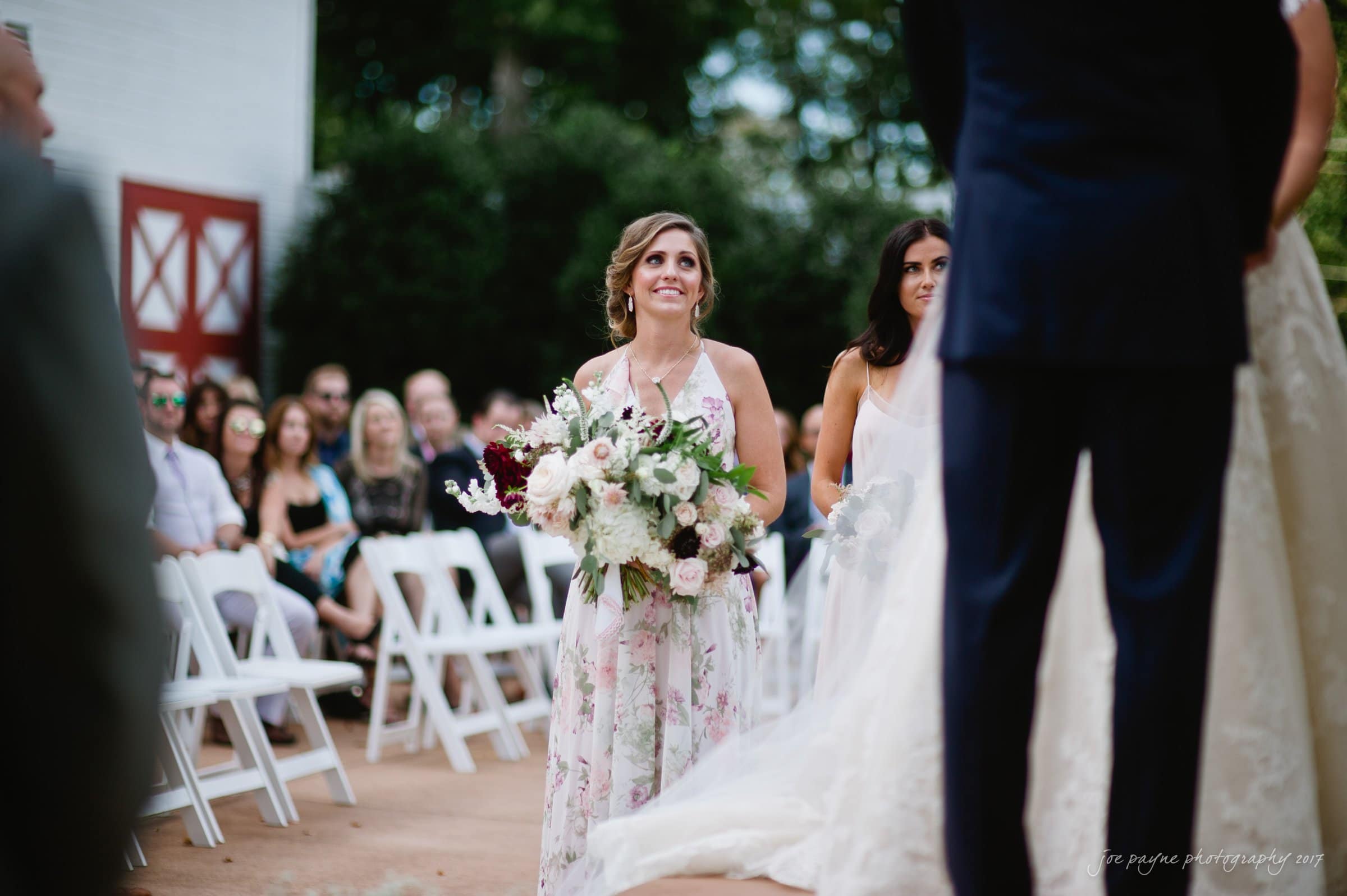 winmock nc wedding photographer - hallie & cullen A woman in a floral dress holds a large bouquet, standing at an outdoor wedding ceremony. Guests are seated on white chairs, watching from behind her. The focus is on the bridal party, with greenery and a barn in the background.