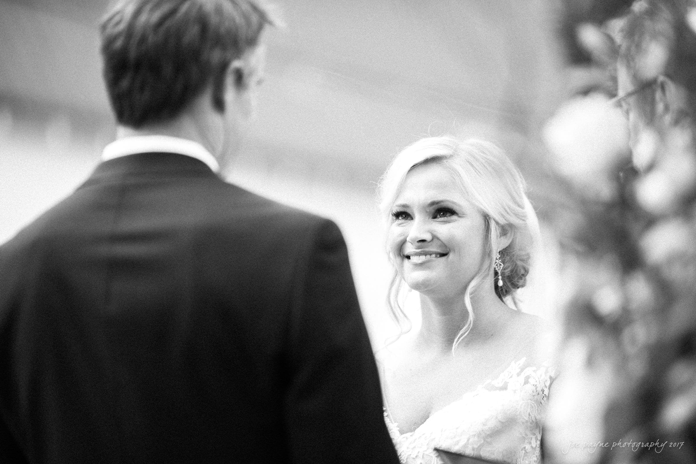 winmock nc wedding photographer - hallie & cullen A black and white photo of a bride and groom during their wedding ceremony. The bride, smiling, looks at the groom as they stand closely. The groom faces her with his back to the camera. The setting has floral decorations.