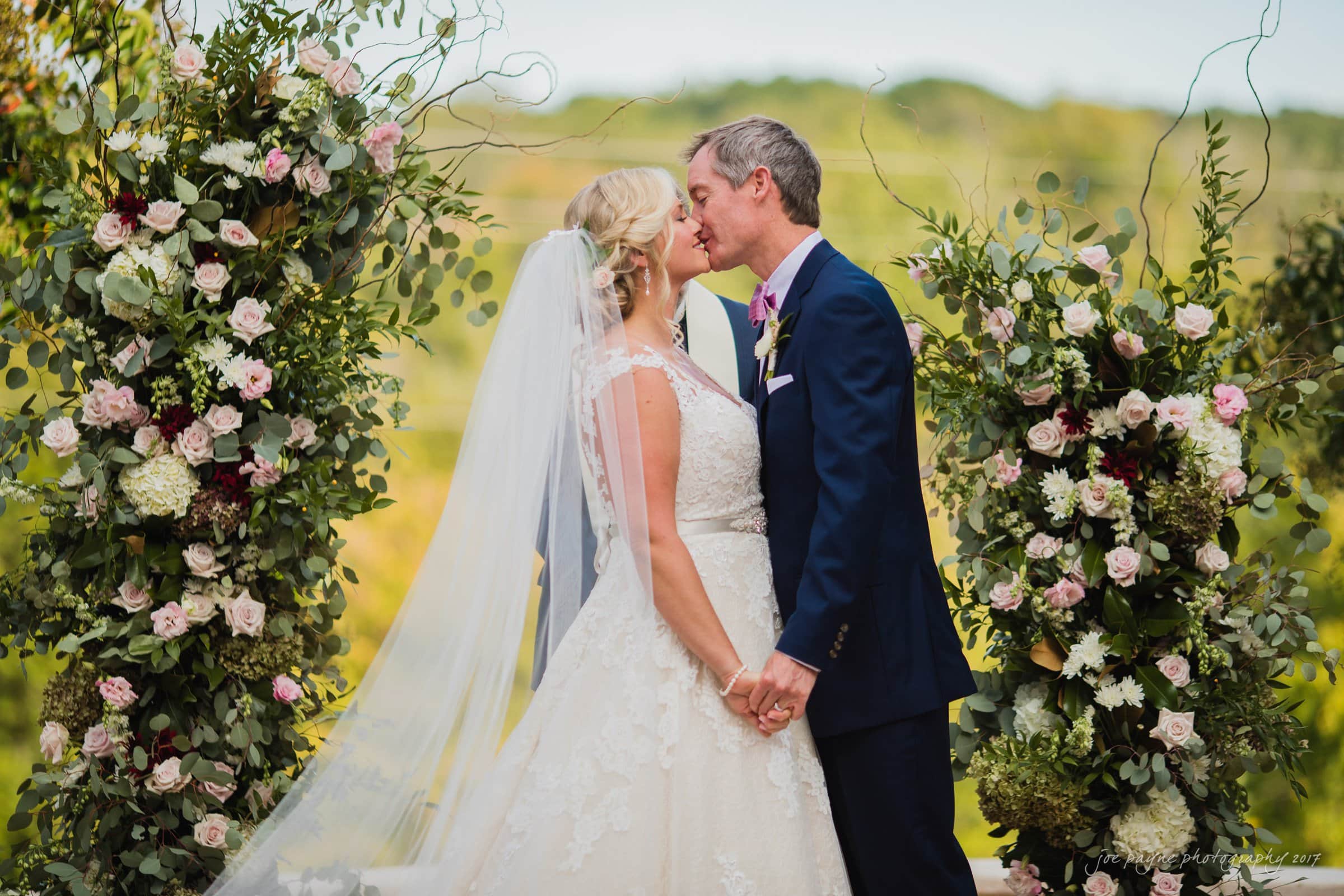 winmock nc wedding photographer - hallie & cullen A bride and groom share a kiss outdoors in front of lush floral arrangements. The bride wears a white gown and veil, while the groom is in a dark suit. The background features greenery and a softly blurred landscape, creating a romantic setting.