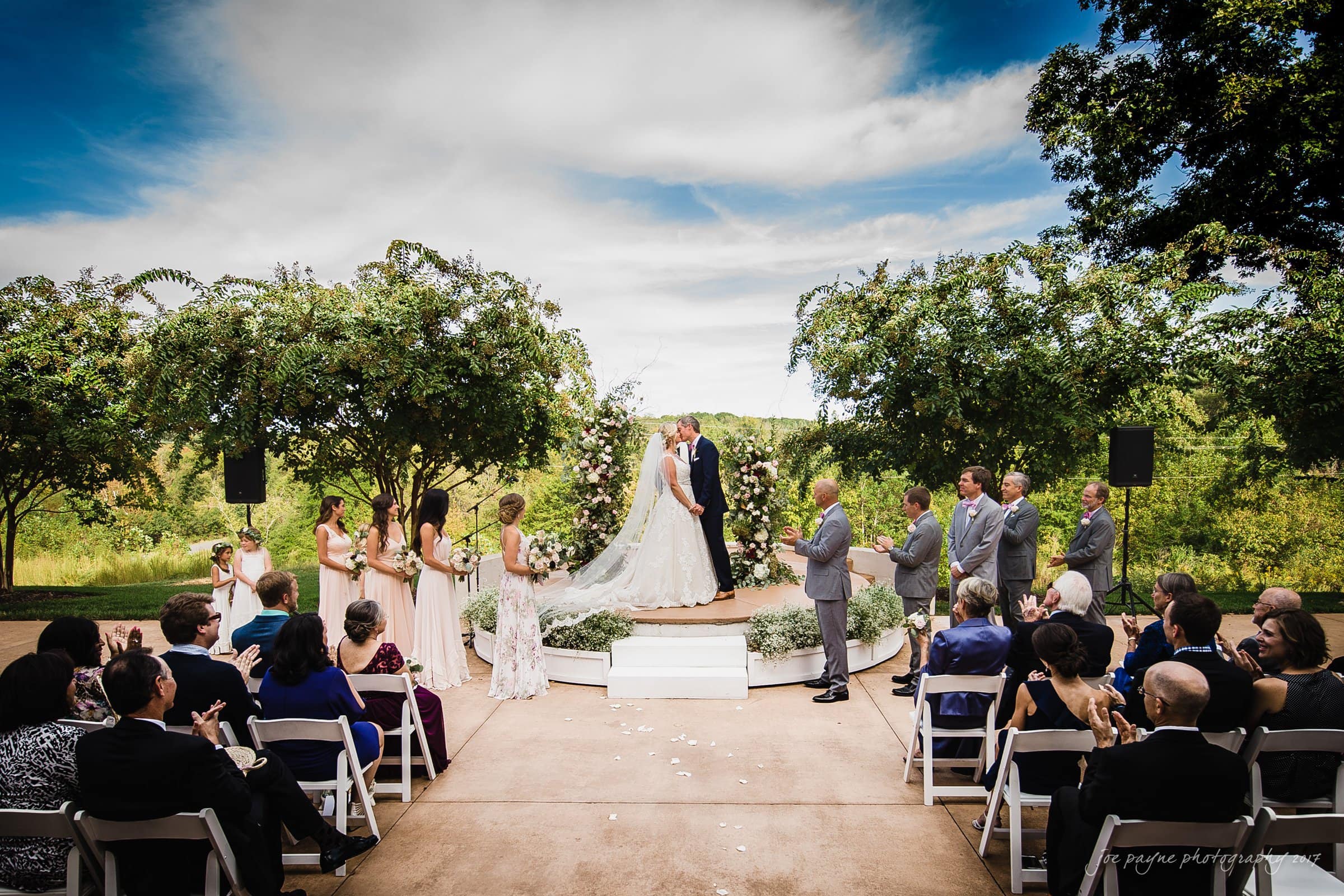 winmock nc wedding photographer - hallie & cullen A bride and groom share a kiss on an outdoor circular platform surrounded by trees. Bridesmaids in light dresses and groomsmen in gray suits stand by. Guests in seated rows watch the ceremony under a bright, partly cloudy sky.