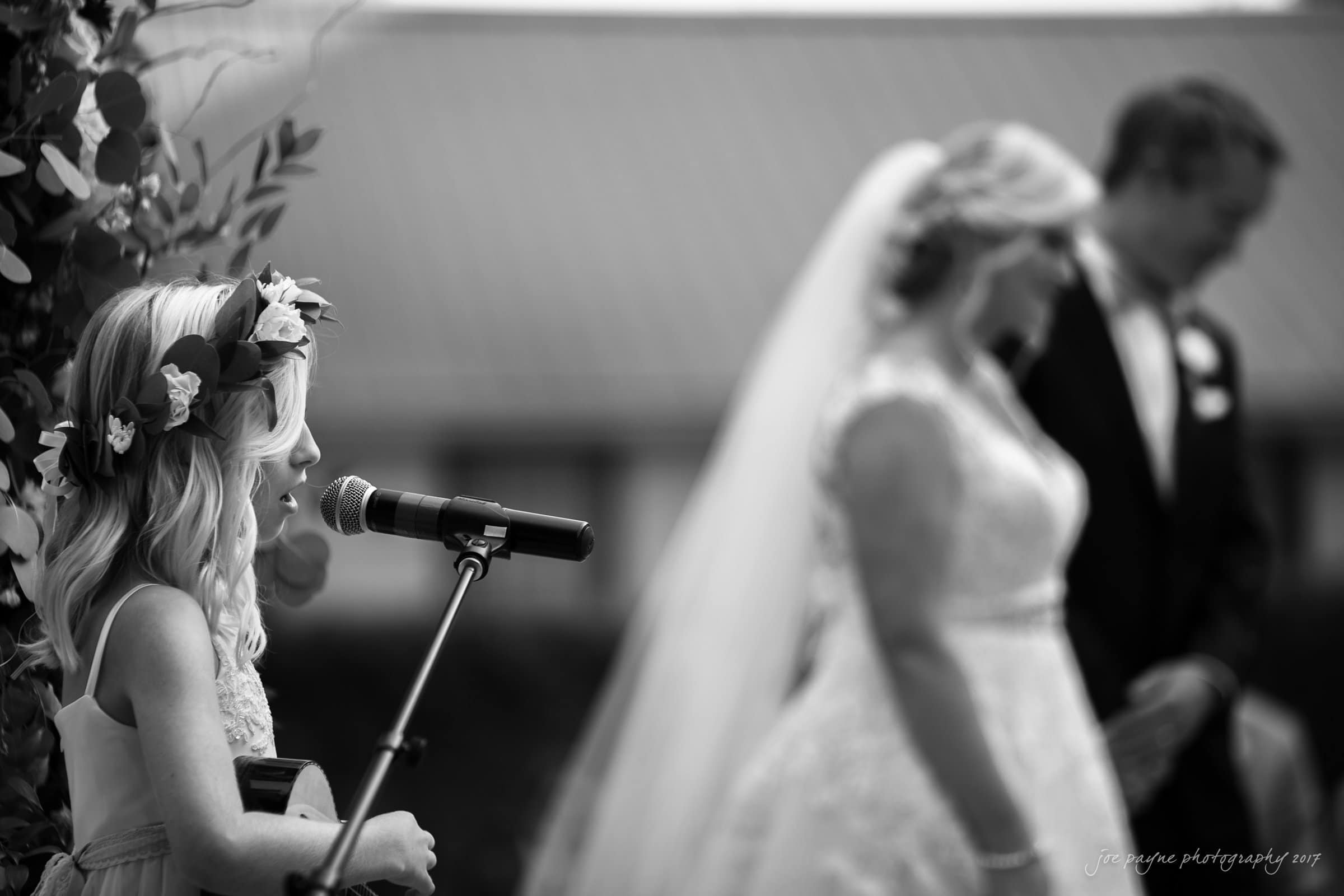 winmock nc wedding photographer - hallie & cullen A young girl with a flower crown sings into a microphone at a wedding ceremony. In the background, a bride in a veil and groom stand together, slightly out of focus. The scene is in black and white.