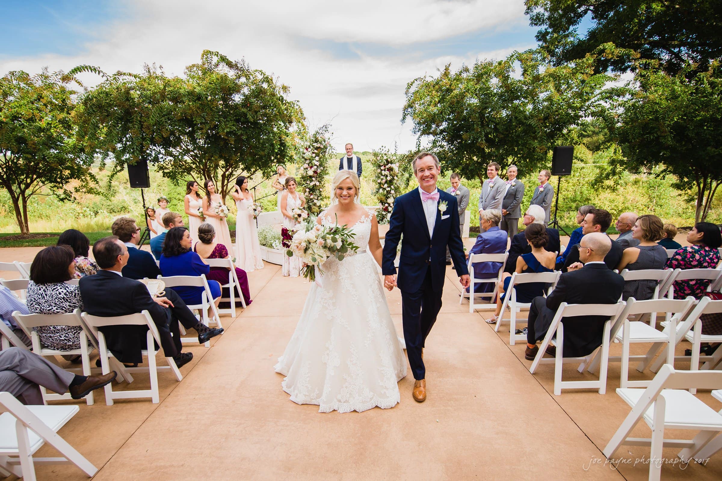 winmock nc wedding photographer - hallie & cullen A newlywed couple walks hand in hand down an outdoor aisle, surrounded by seated guests. The bride wears a white gown and holds a bouquet, while the groom wears a navy suit with a pink bow tie. Bridesmaids and a groomsman stand under trees in the background.