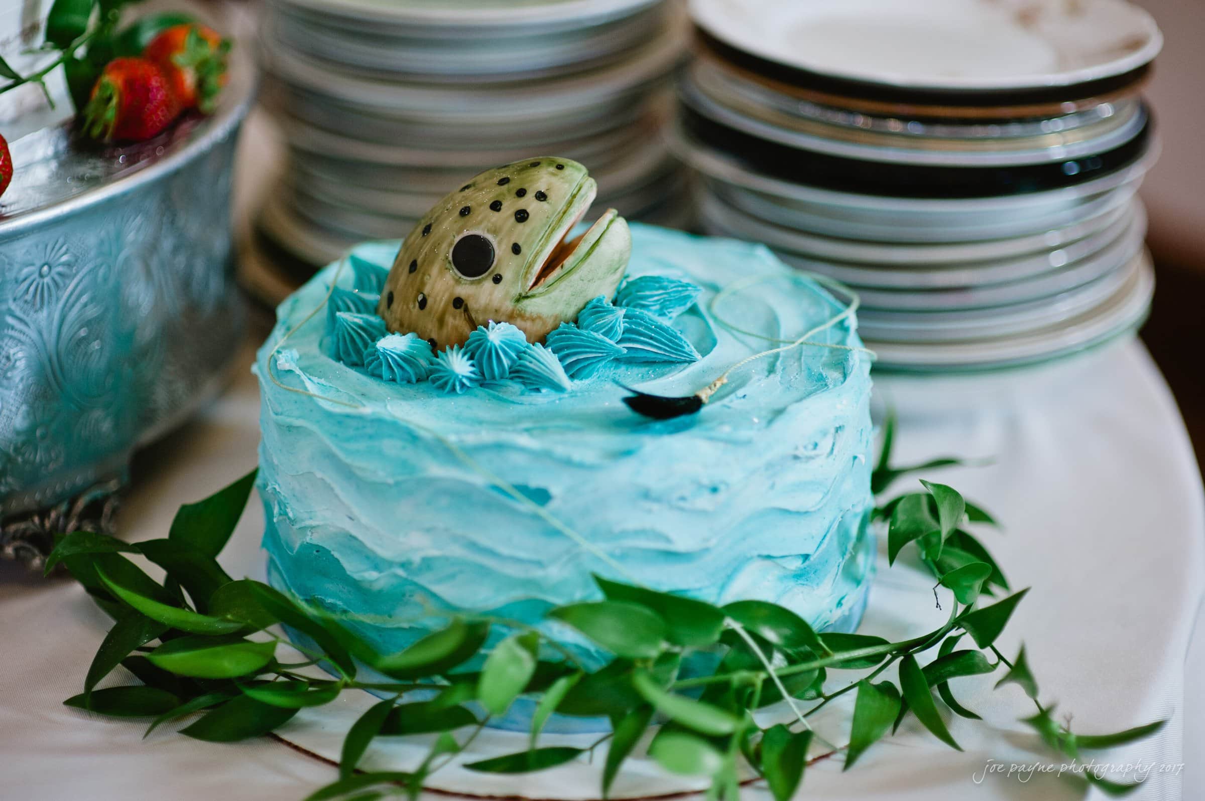 winmock nc wedding photographer - hallie & cullen A novelty cake with blue frosting, resembling waves, features a realistic fish head protruding from its top. Its surrounded by green leaves and sits next to a stack of plates on a white tablecloth.