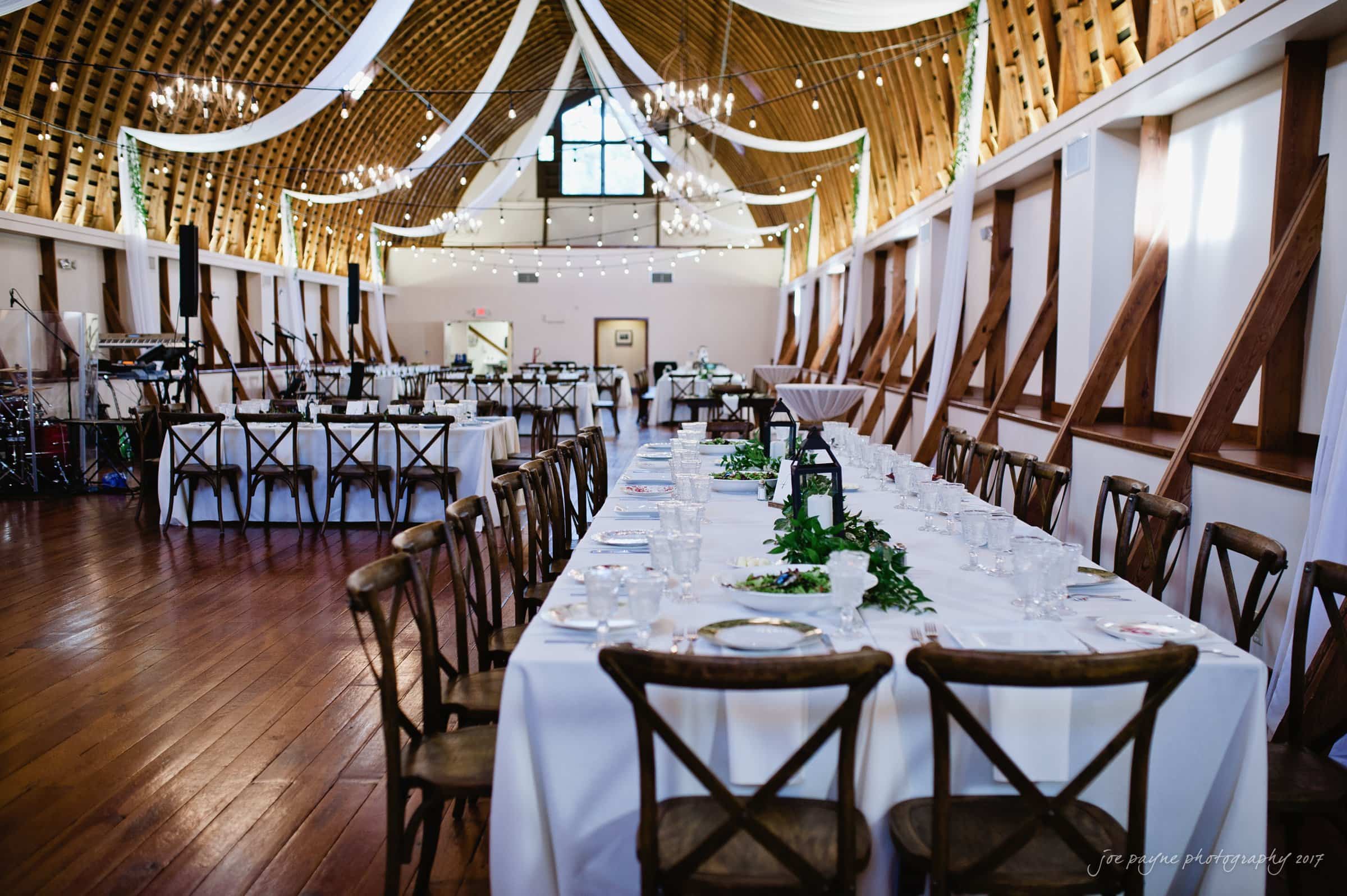 winmock nc wedding photographer - hallie & cullen A rustic wedding reception setup inside a barn with long tables covered in white tablecloths, wooden cross-back chairs, and elegant chandeliers hanging from the wooden ceiling. White drapes and greenery add to the decor.