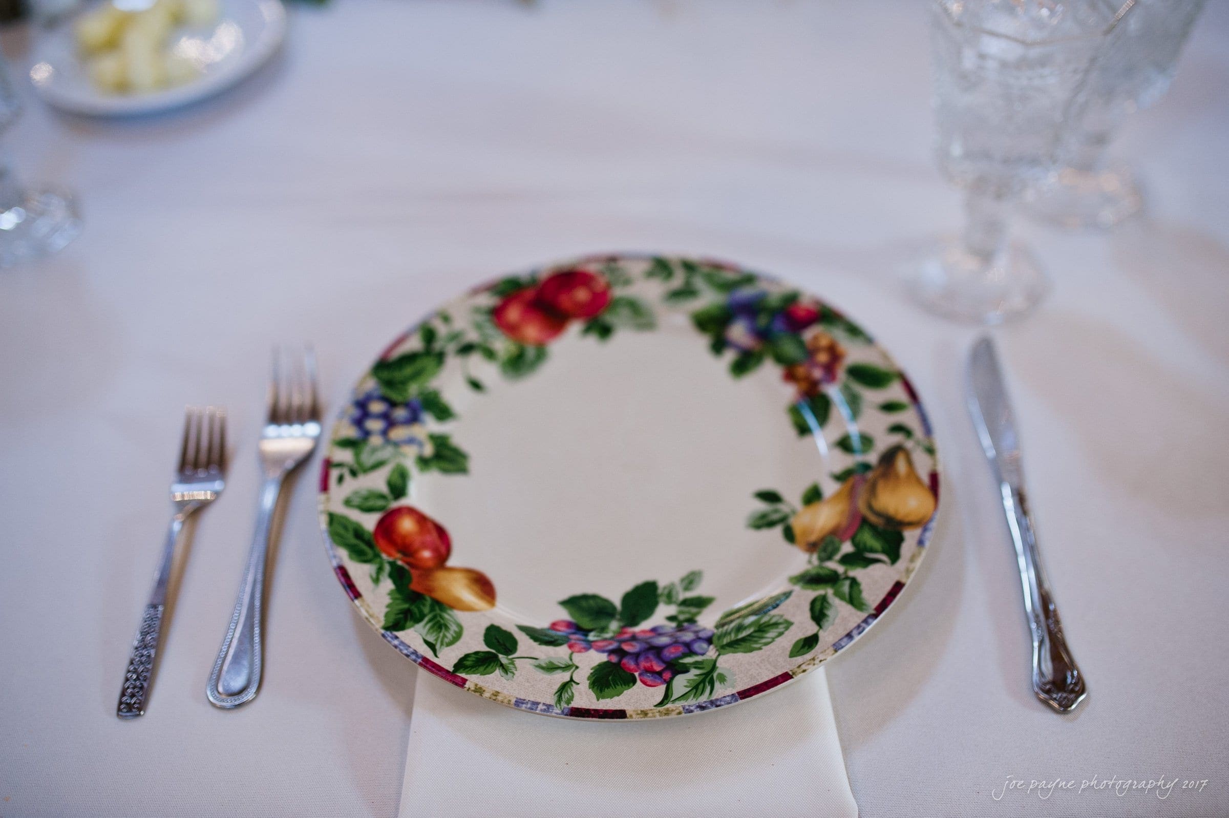winmock nc wedding photographer - hallie & cullen A decorative table setting with a floral-patterned plate centered on a white tablecloth. The plate is flanked by a fork on the left, a knife and spoon on the right, and a folded napkin beneath. Crystal glassware is positioned in the background.