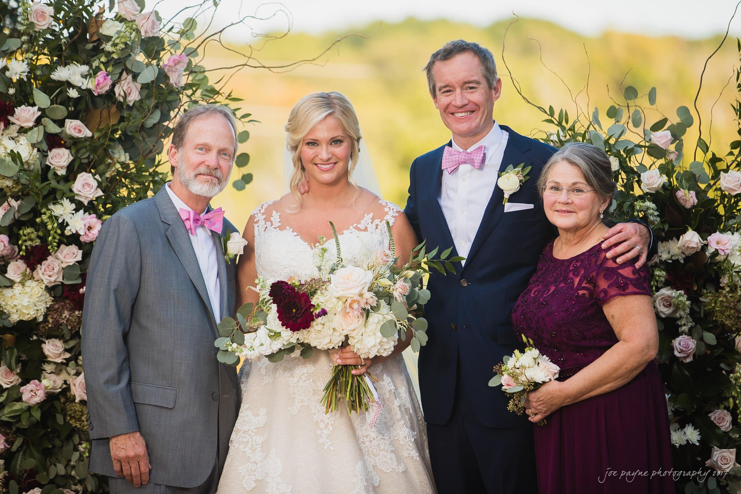 winmock nc wedding photographer - hallie & cullen A bride and groom stand outdoors with an older couple. The bride holds a bouquet, wearing a white gown, and the groom wears a dark suit with a bow tie. The older man is in a gray suit, and the older woman wears a purple dress. Floral arrangements surround them.