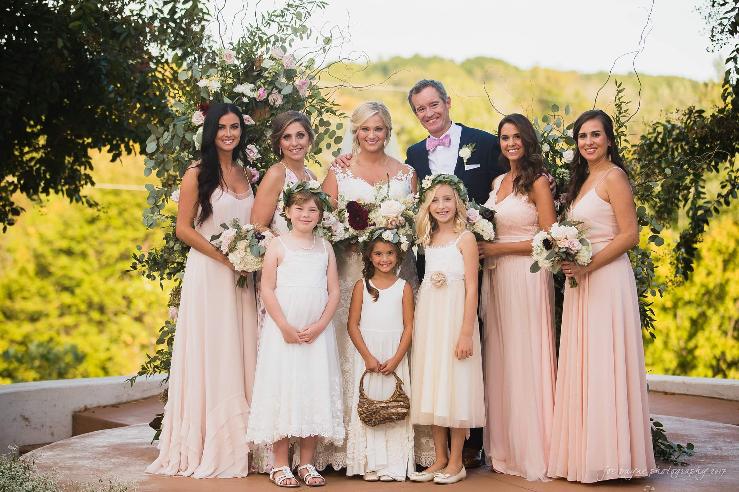 winmock nc wedding photographer - hallie & cullen A wedding party stands outdoors, with a bride in the center holding a bouquet, surrounded by bridesmaids in light pink dresses, and a groom in a suit and pink bowtie. Two flower girls stand in front, holding baskets. Lush greenery forms the backdrop.