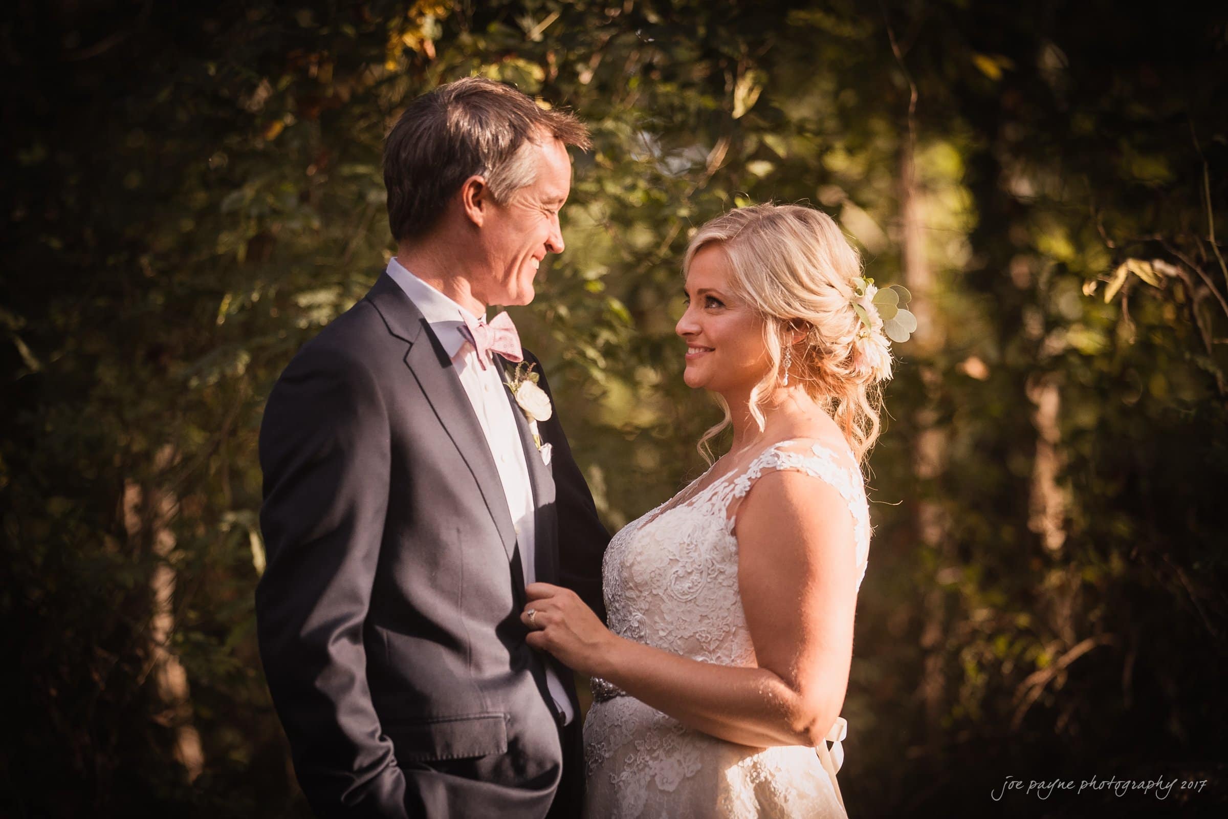 winmock nc wedding photographer - hallie & cullen A couple stands facing each other in a sunlit forest setting. The man wears a dark suit with a bow tie, and the woman is in a white lace dress with flowers in her hair. They smile warmly at each other.