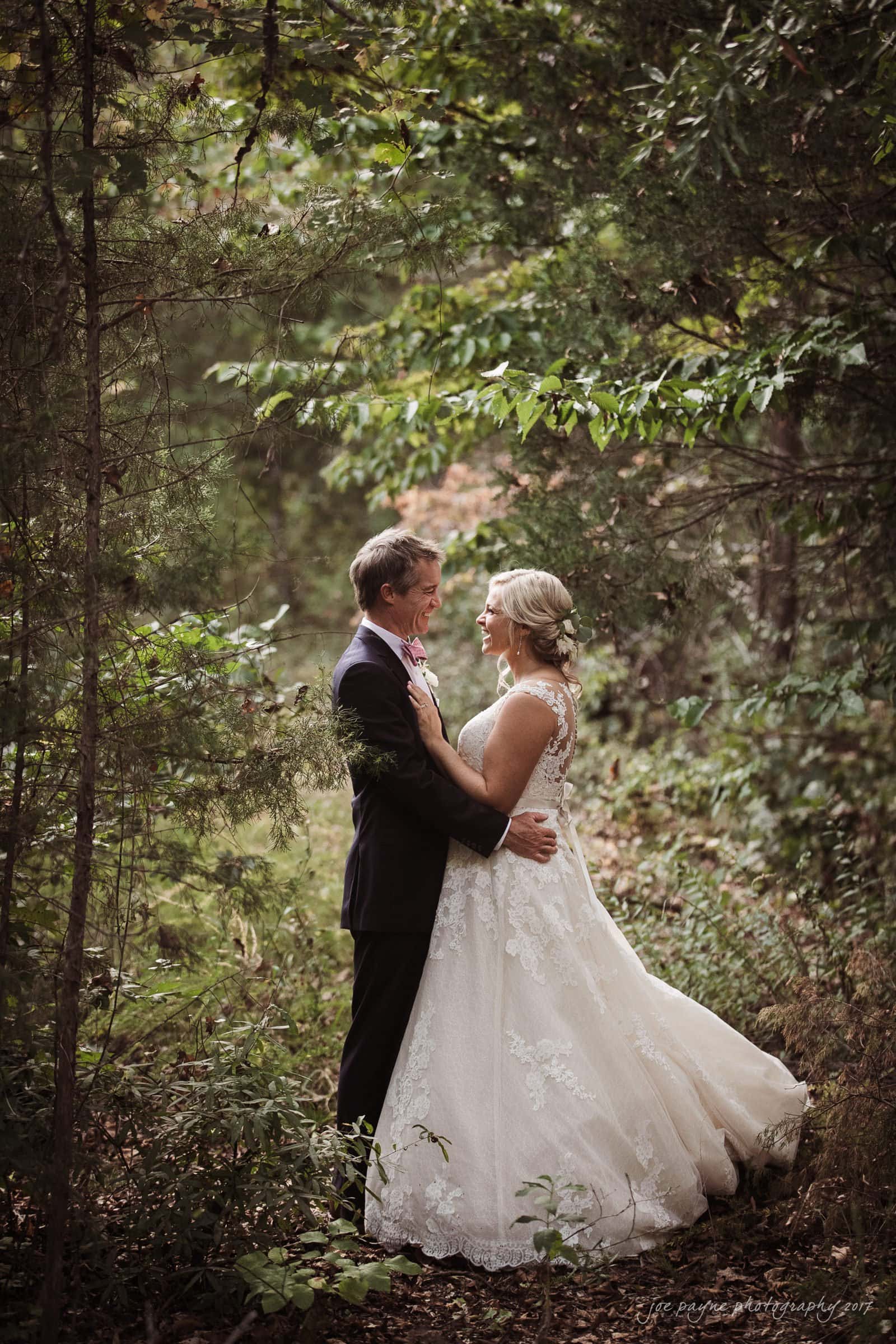 winmock nc wedding photographer - hallie & cullen A bride and groom stand in a lush forest, gazing into each others eyes. The bride is wearing a white lace gown, and the groom is in a dark suit. Sunlight filters through the trees, creating a romantic atmosphere.