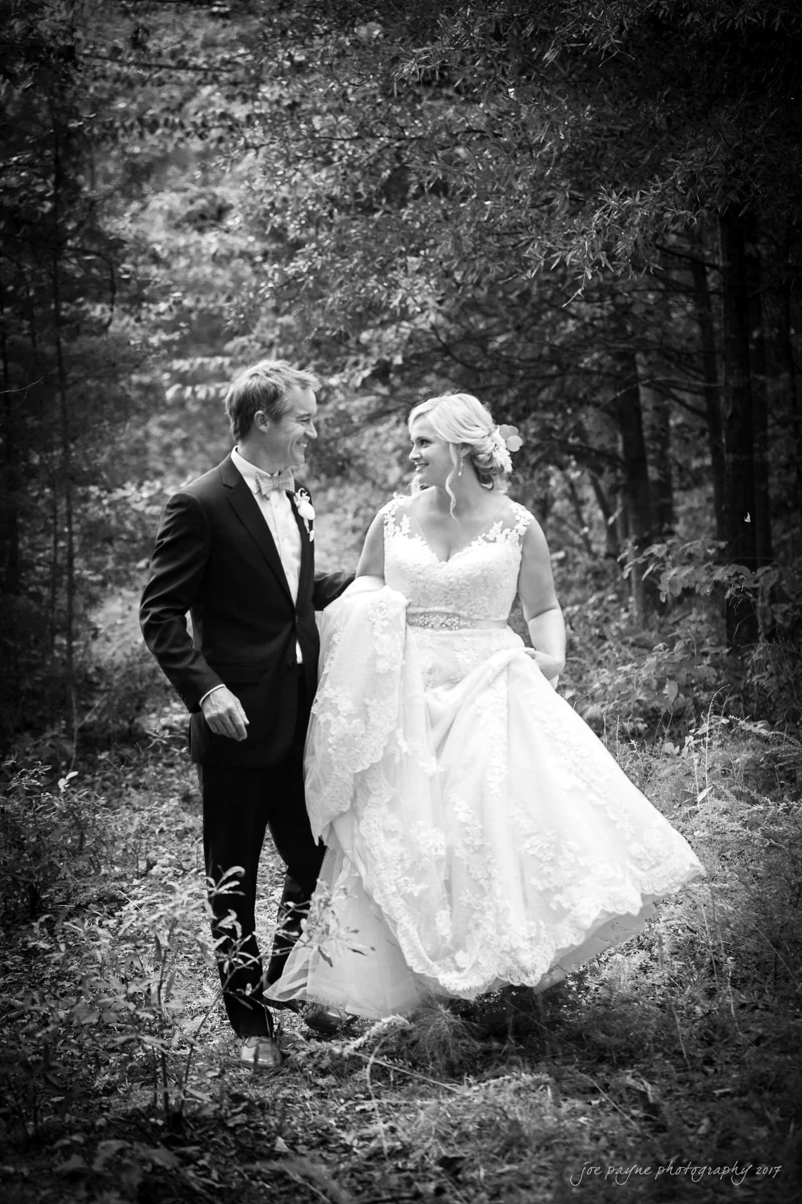 winmock nc wedding photographer - hallie & cullen A bride and groom stand in a wooded area, gazing at each other. The bride wears a lace wedding dress with a train, while the groom is in a suit. The black-and-white photo captures a serene and romantic moment.