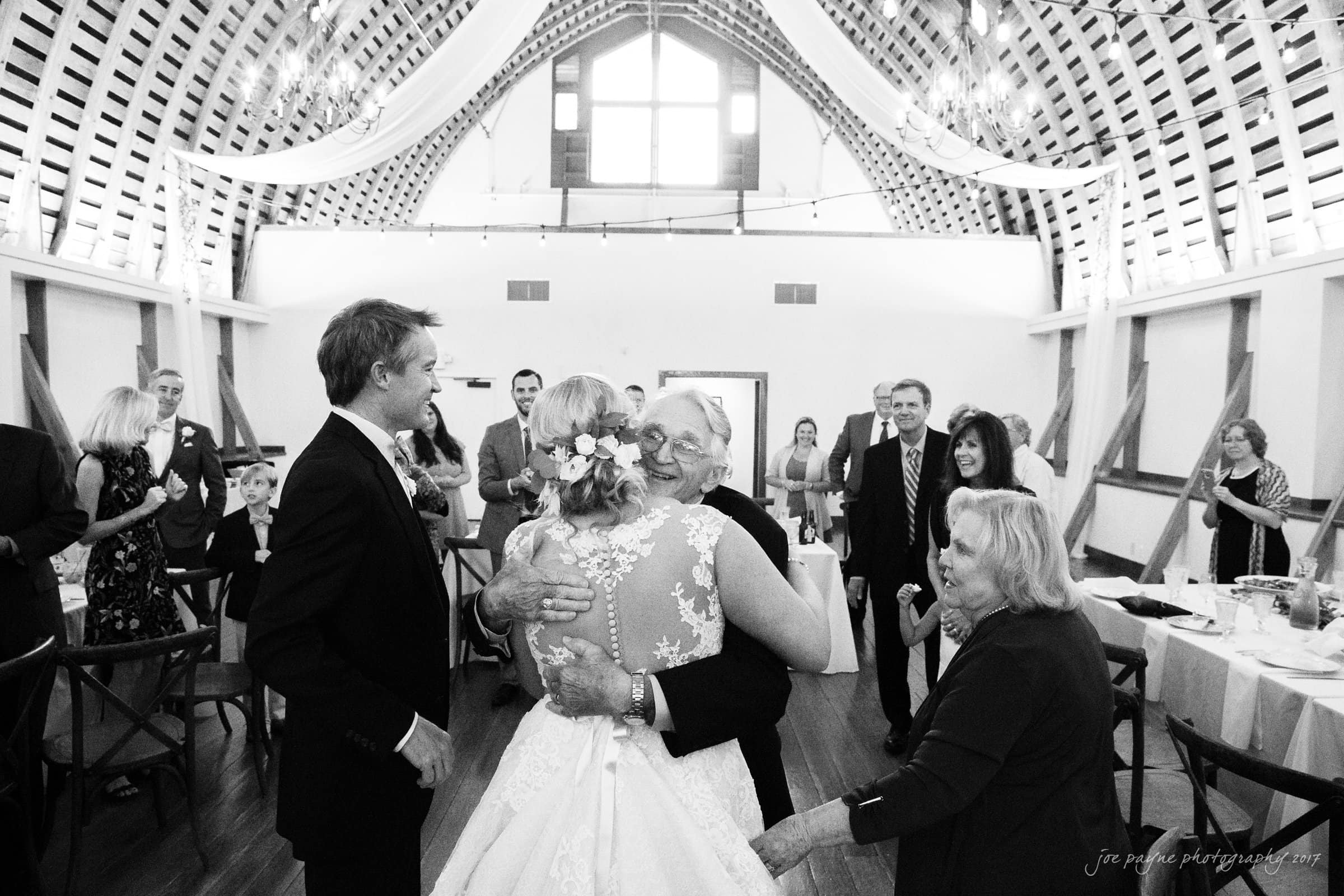 winmock nc wedding photographer - hallie & cullen A bride in a lace dress hugs an elderly man at a wedding reception inside a barn-style venue. Guests stand around smiling and clapping. The ceiling features draped fabric and string lights, creating a warm and festive atmosphere.
