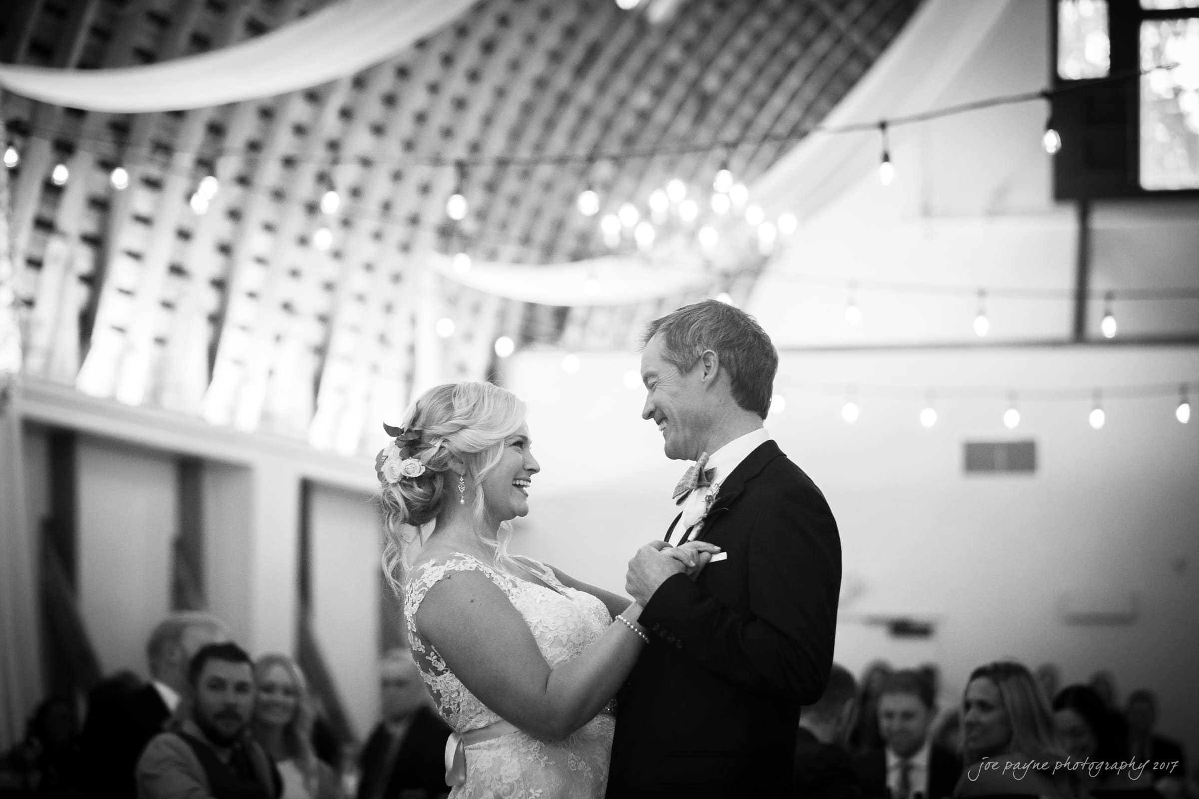 winmock nc wedding photographer - hallie & cullen A couple shares a joyful dance at a wedding reception in a decorated hall, with draped fabric and string lights hanging from the curved ceiling. Guests are seated in the background, watching and smiling.