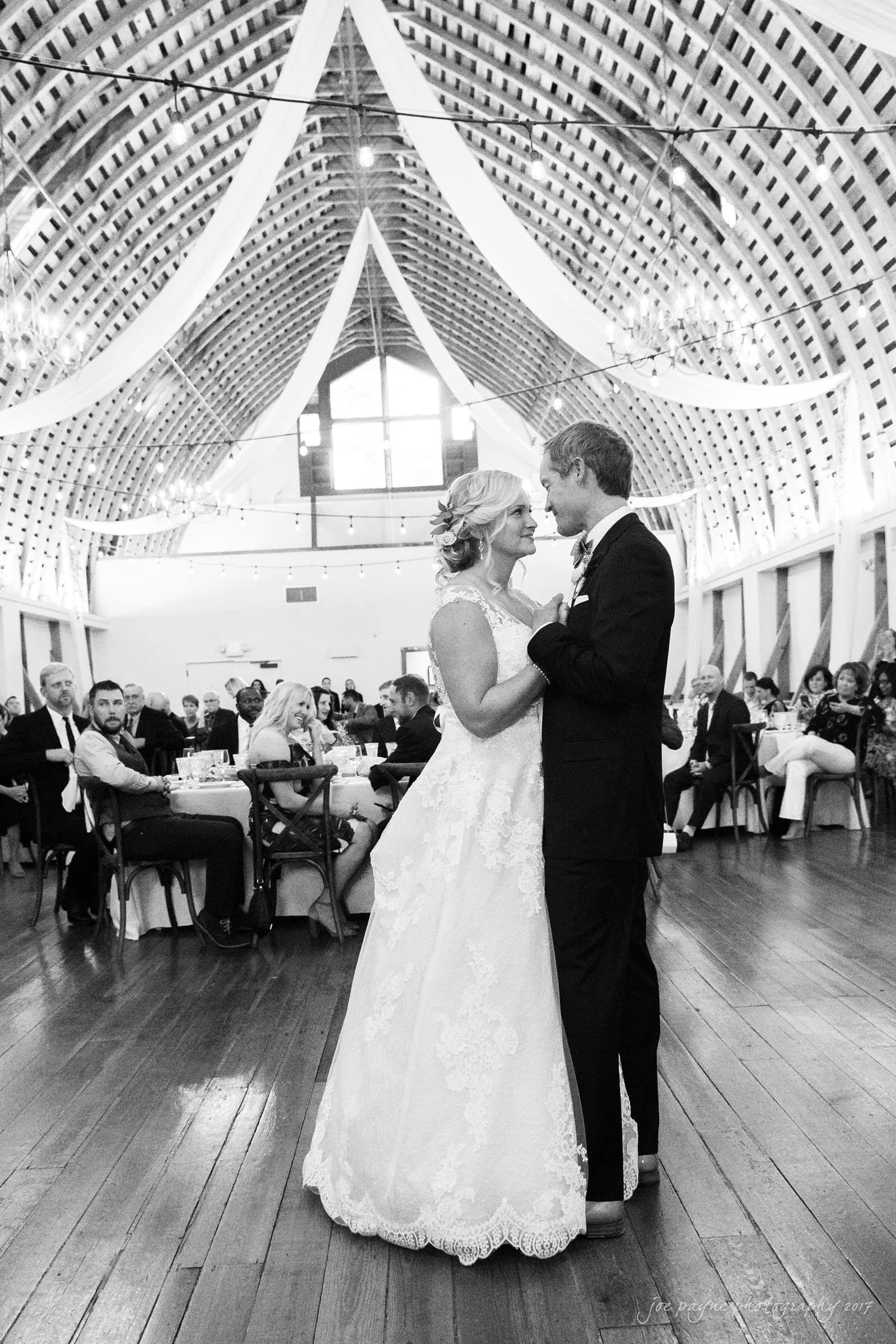 winmock nc wedding photographer - hallie & cullen A bride and groom share their first dance in a beautifully decorated, spacious hall. Guests sit at round tables, watching the couple. The ceiling features draped fabric and string lights, creating an elegant and romantic atmosphere.