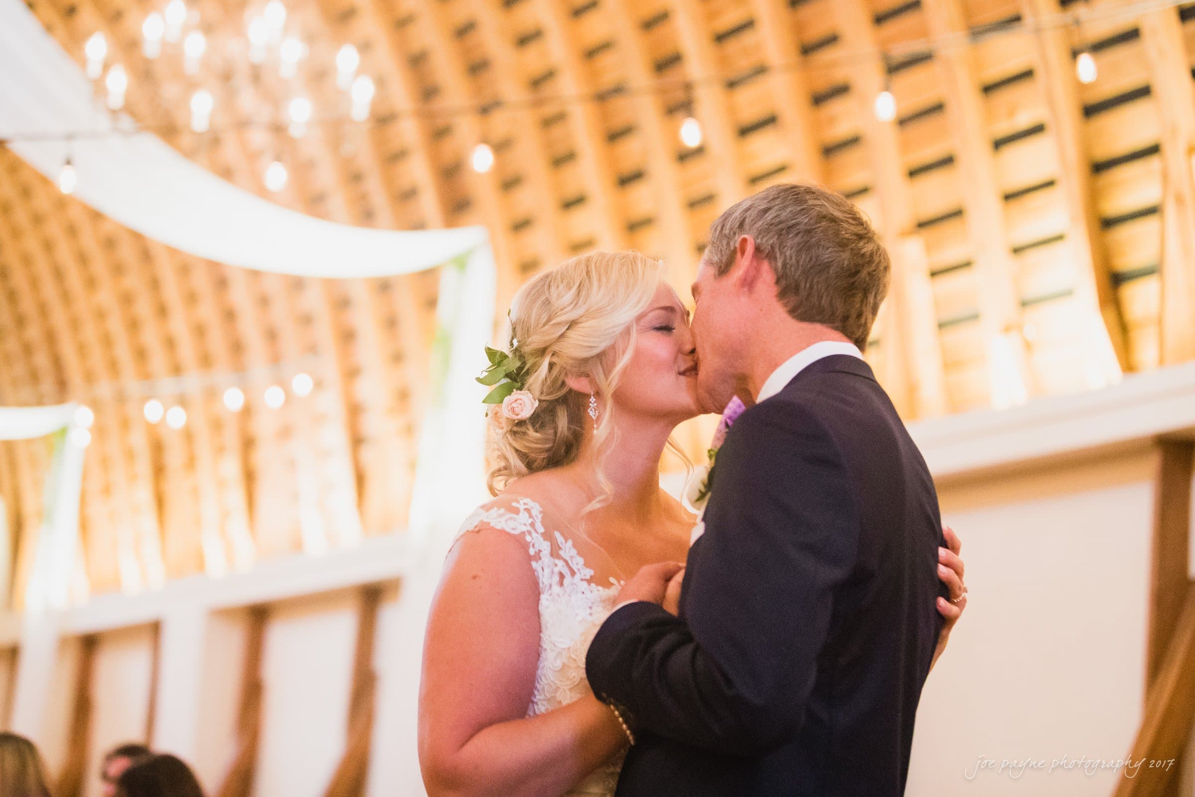 winmock nc wedding photographer - hallie & cullen A bride and groom share a kiss during their wedding dance in a warmly lit barn with string lights. The bride wears a lace gown and a floral hairpiece, while the groom is in a dark suit. The atmosphere is romantic and intimate.