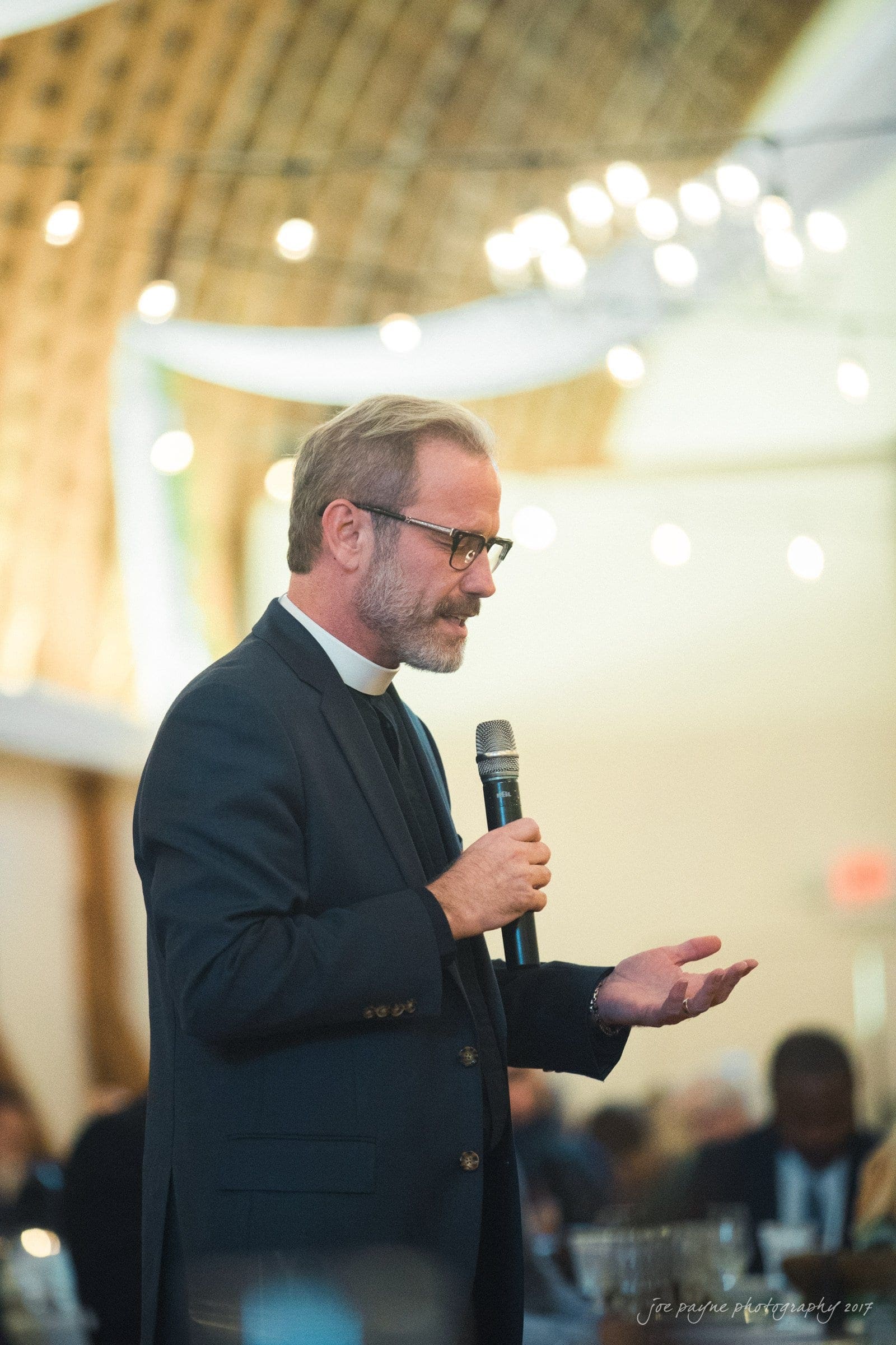 winmock nc wedding photographer - hallie & cullen A man in a suit and clerical collar speaks into a microphone at an indoor event. The setting is warmly lit with string lights and a blurred audience in the background.