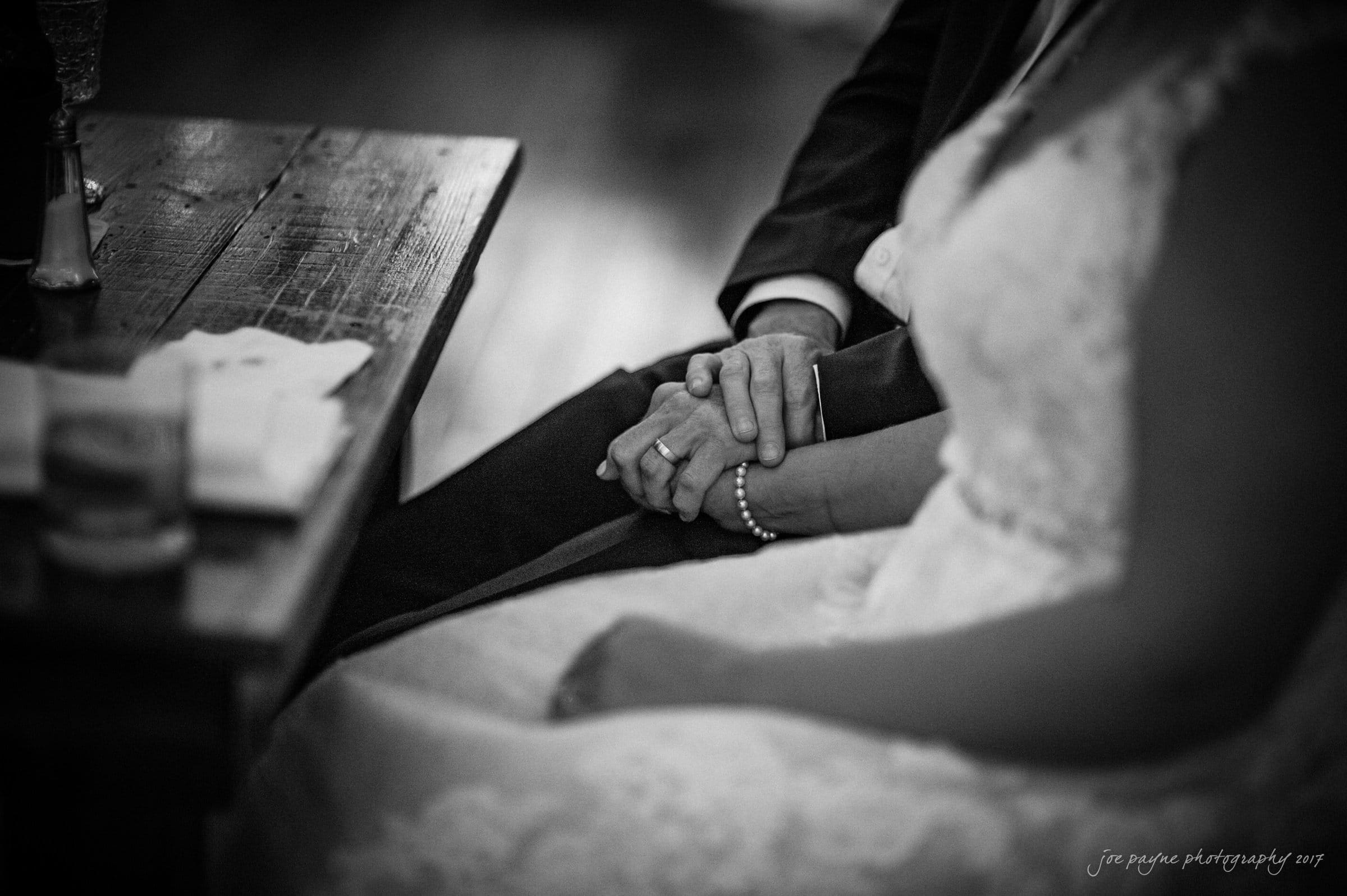 winmock nc wedding photographer - hallie & cullen A bride and groom sit closely at a wooden table, holding hands. The bride is in a lace dress, and the groom is in a suit. The image is in black and white, capturing an intimate moment of togetherness.