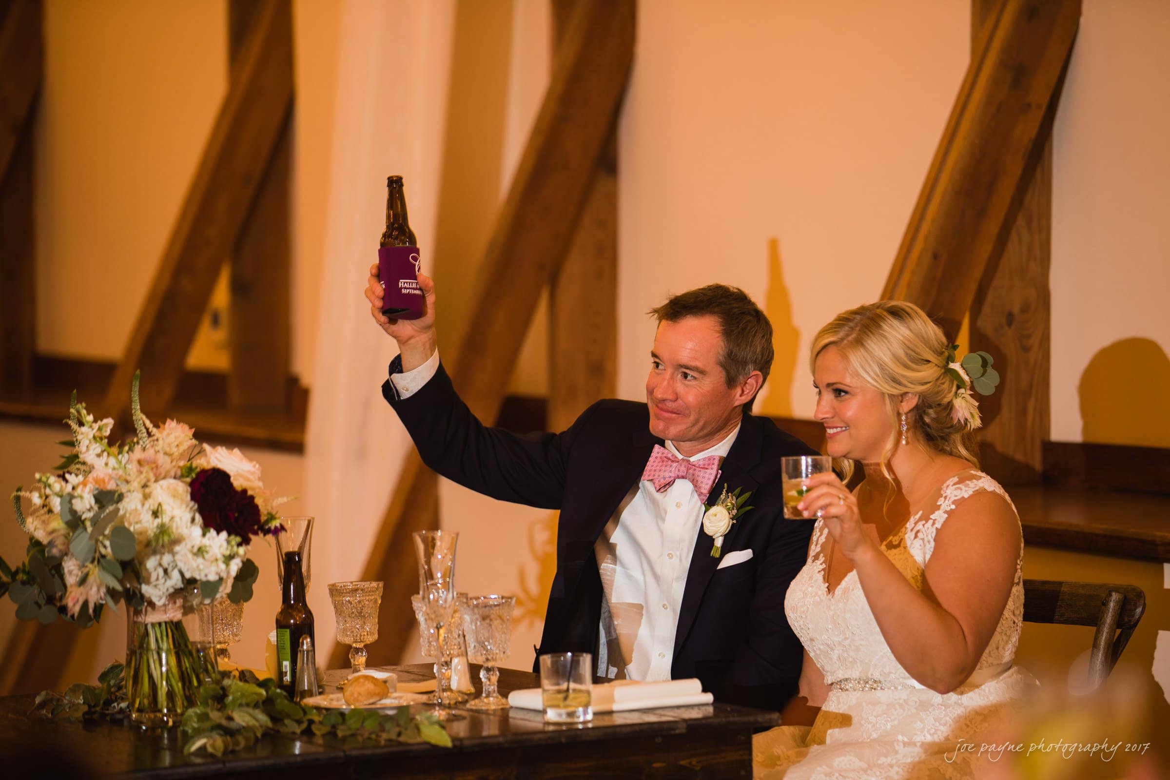 winmock nc wedding photographer - hallie & cullen A bride and groom sit together at a table during their wedding reception. The groom raises a beer bottle in celebration, while the bride holds a glass. The table is decorated with floral arrangements and glasses. They are smiling happily.