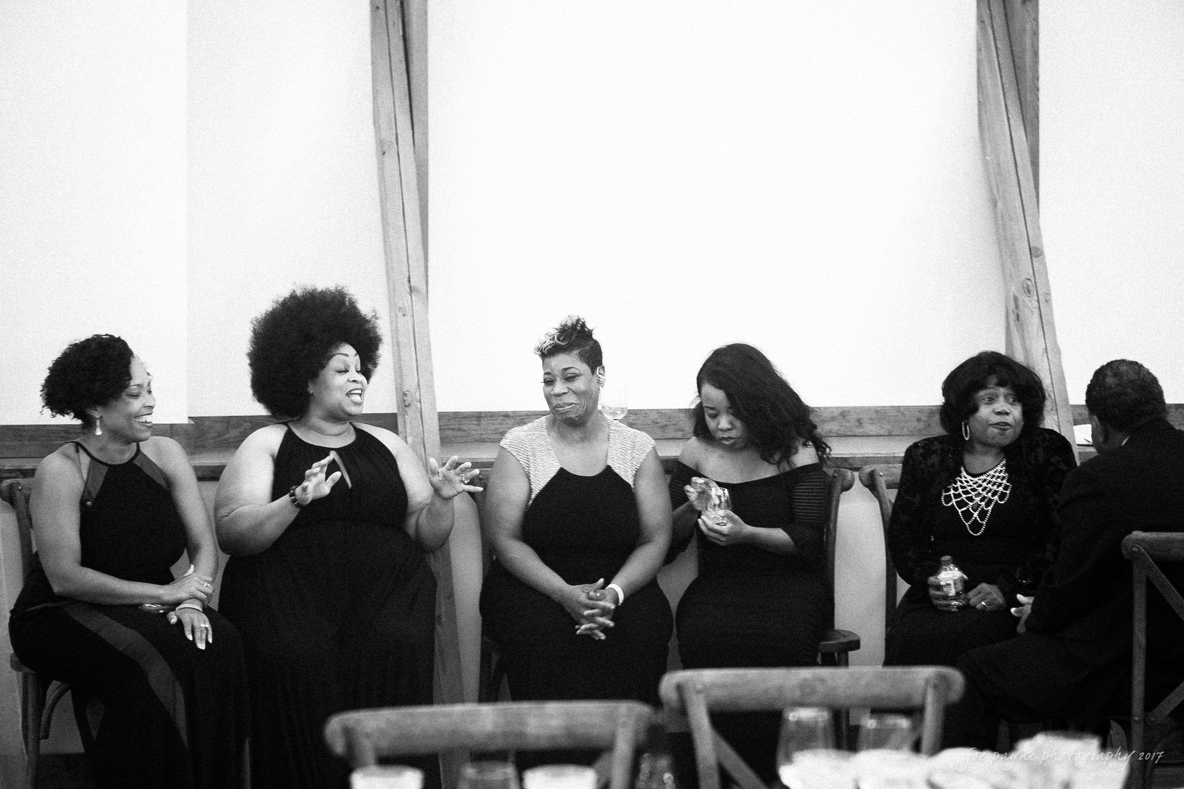 winmock nc wedding photographer - hallie & cullen Five women in elegant black dresses are sitting together and chatting. They appear to be enjoying each others company in a formal setting, possibly at an event or gathering, with wooden beams visible in the background.