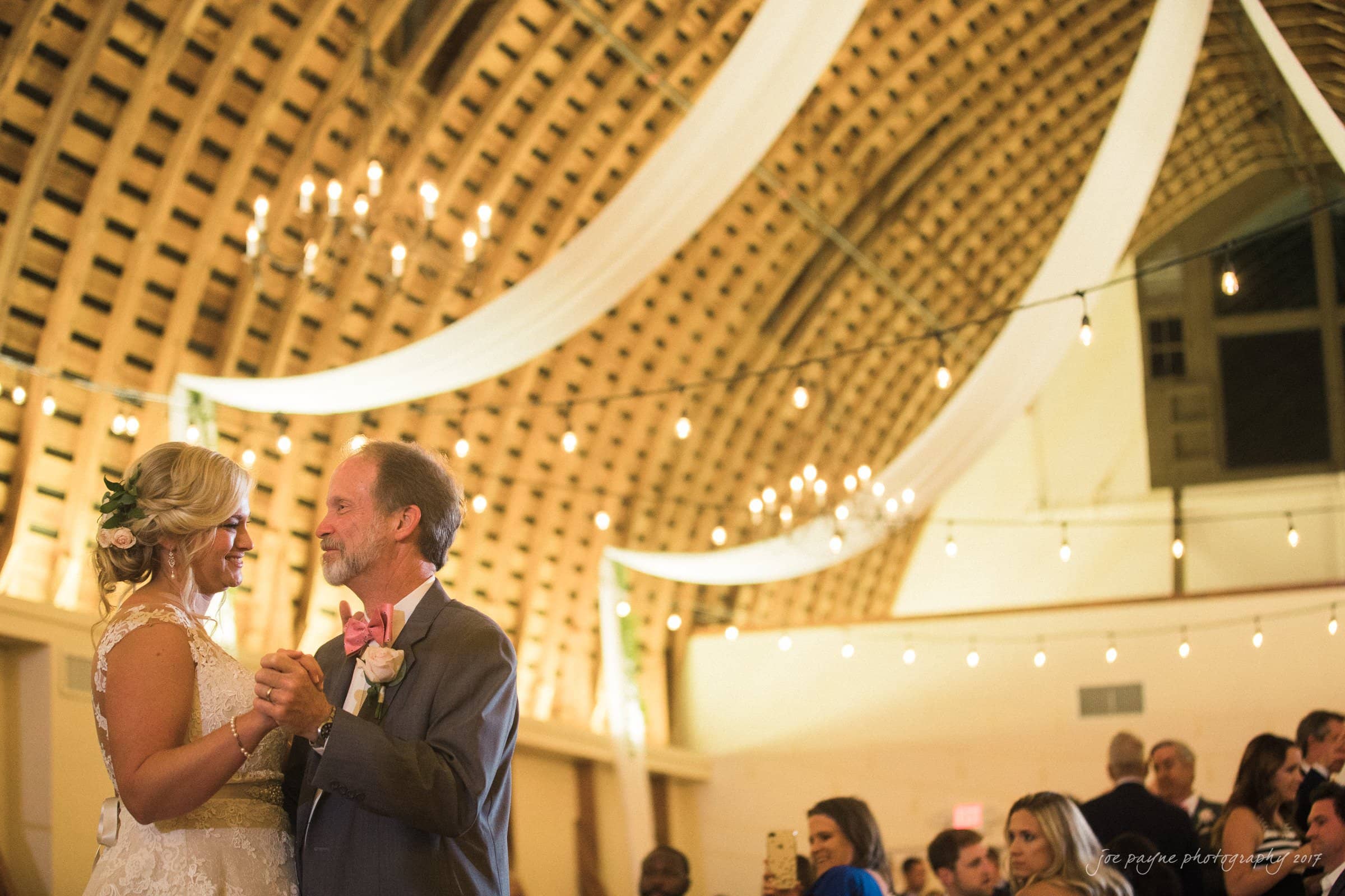 winmock nc wedding photographer - hallie & cullen A bride and groom dance under a canopy of string lights and draped fabric in a warmly lit wooden hall. The bride smiles joyfully at her partner, who is dressed in a suit with a pink bow tie. Guests look on in the background.