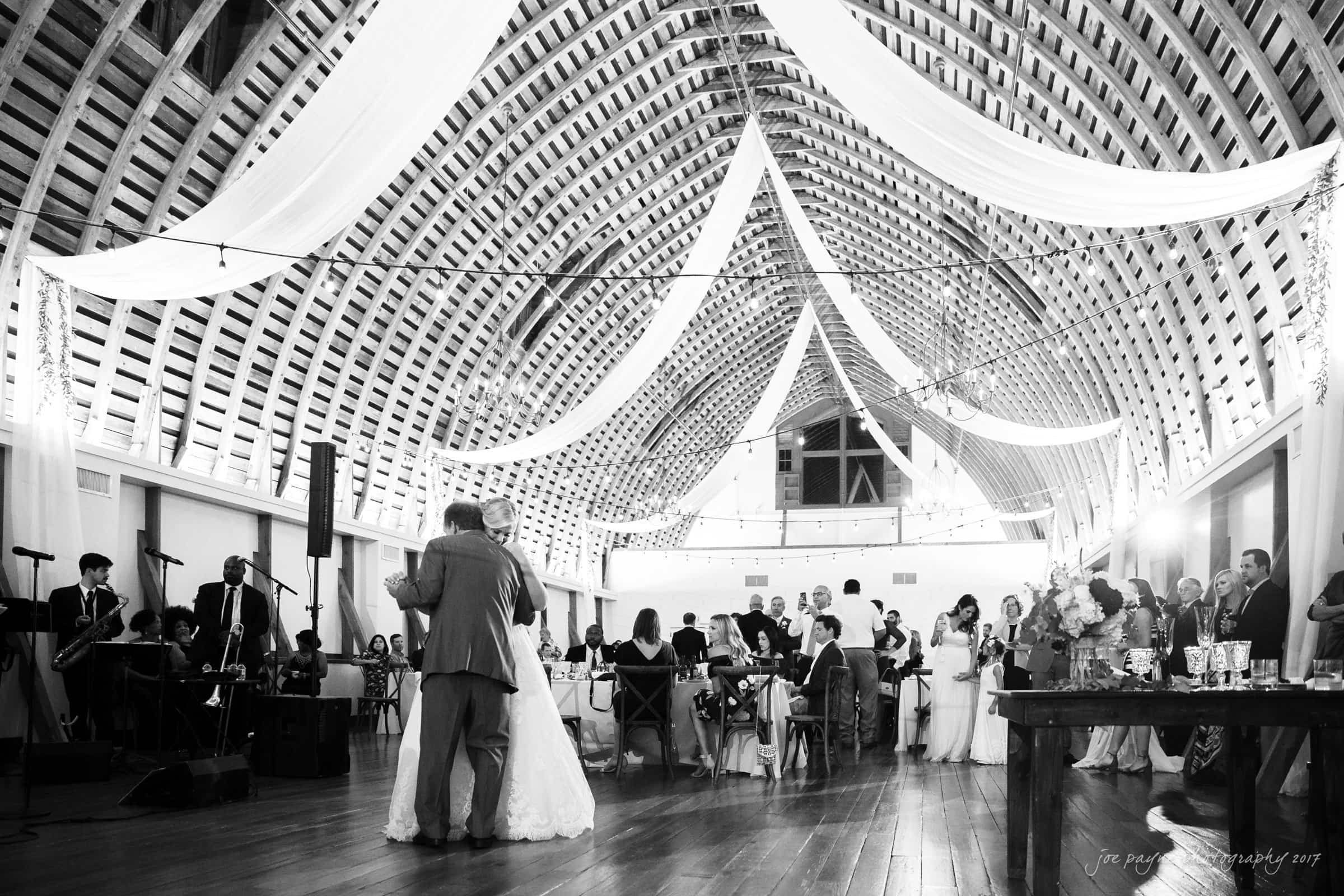 winmock nc wedding photographer - hallie & cullen A newlywed couple shares a first dance in an elegant, rustic barn venue. The ceiling is adorned with draped fabric and string lights. Guests and a live band, dressed formally, observe the couples dance under the ambient lighting.