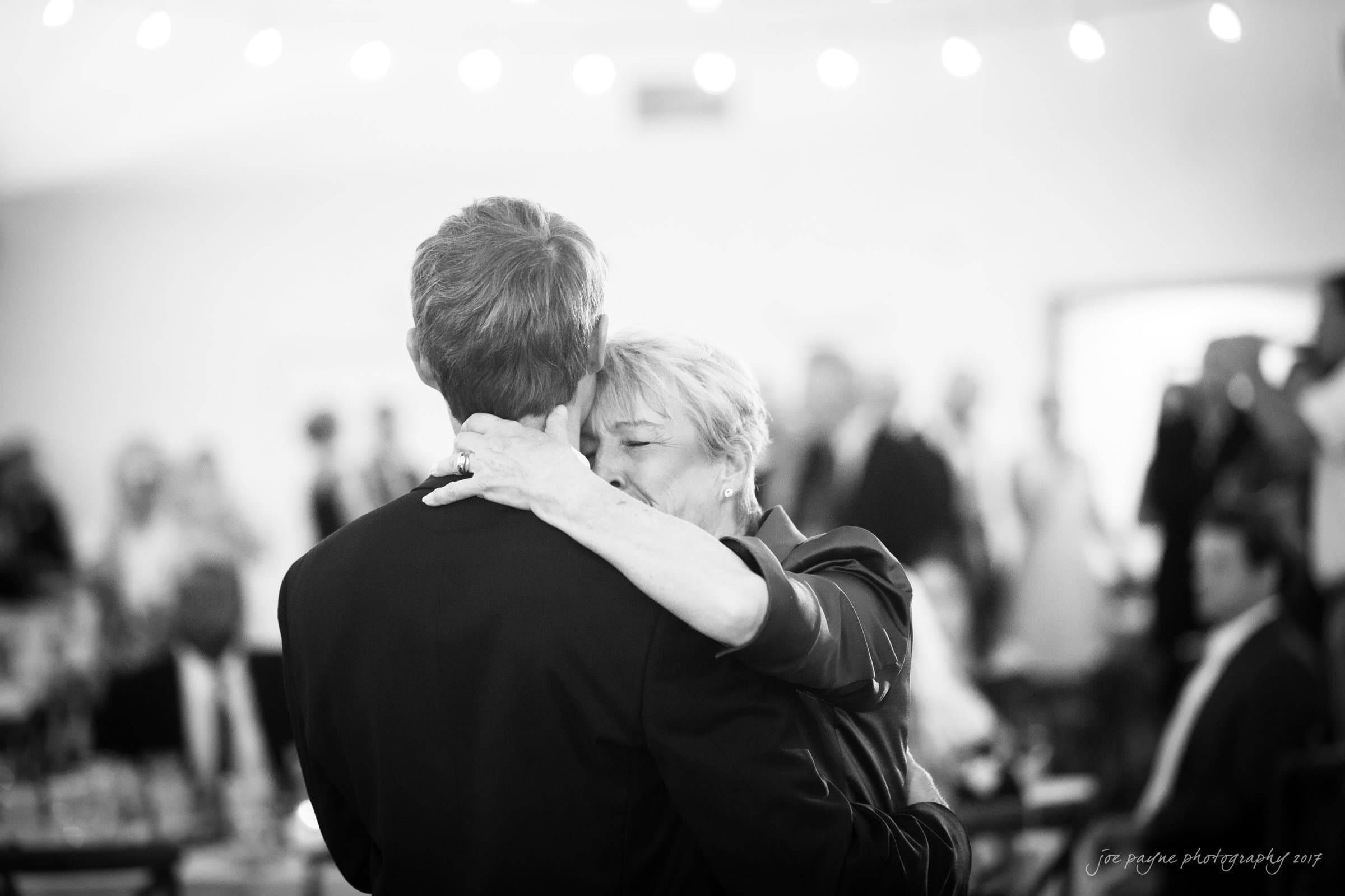 winmock nc wedding photographer - hallie & cullen A black-and-white image of a woman embracing a man, possibly during a dance. The setting seems to be a formal event or celebration, with blurred guests and lights in the background, creating a warm, intimate atmosphere.