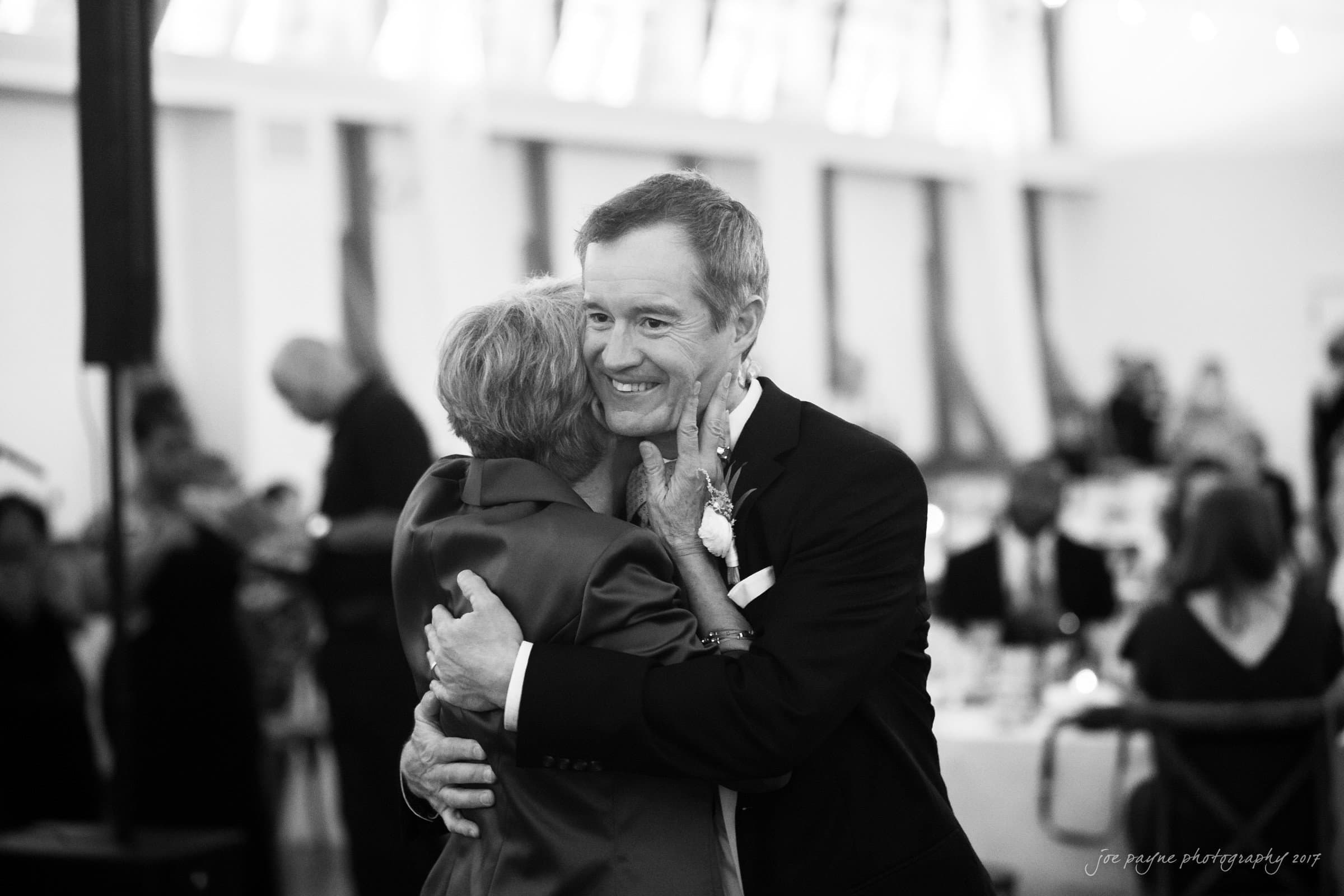 winmock nc wedding photographer - hallie & cullen A black and white photograph of two people embracing warmly on a dance floor. The man is wearing a suit with a boutonniere, and they are surrounded by blurred figures in a softly lit room, suggesting a celebratory event.