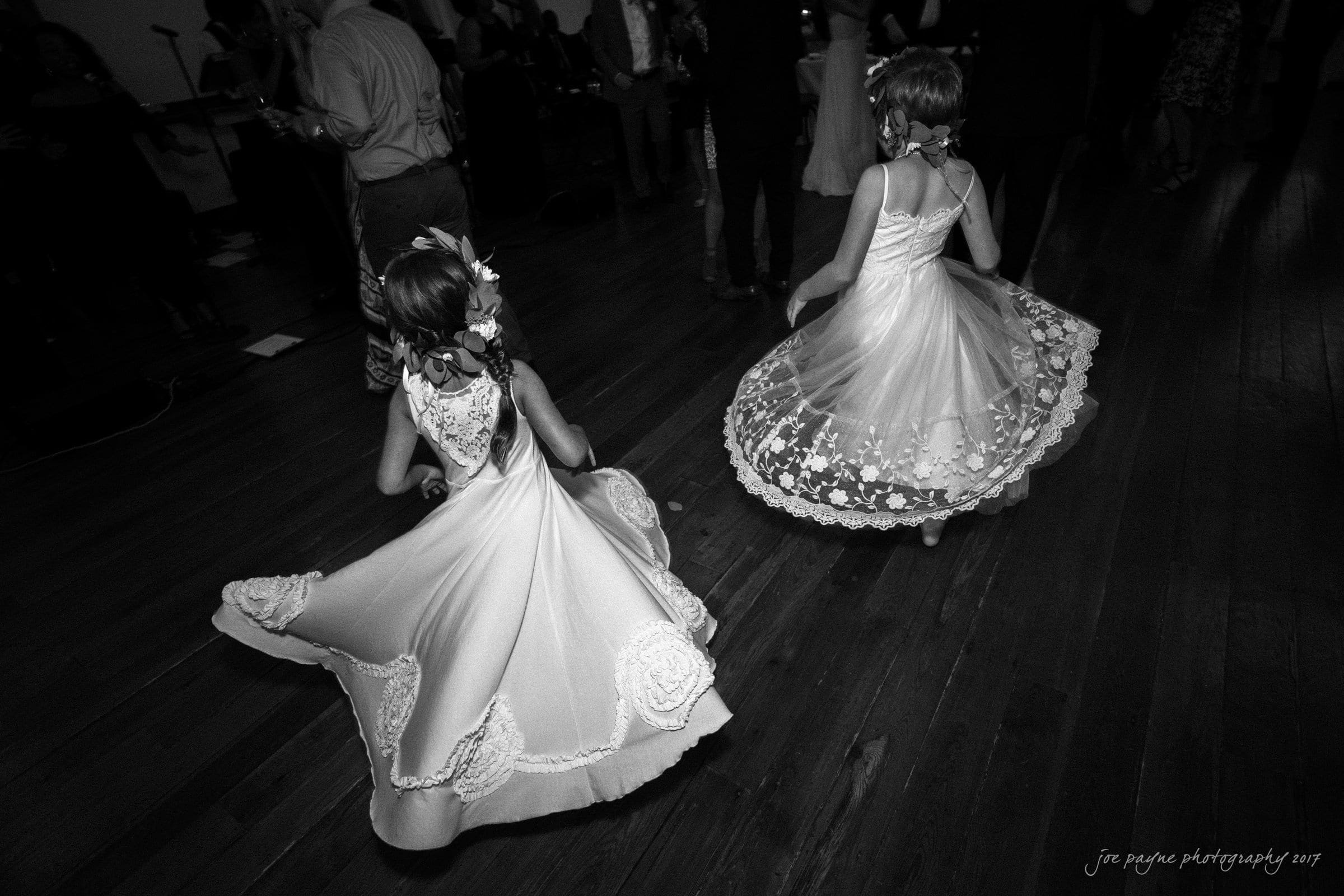 winmock nc wedding photographer - hallie & cullen Two young girls in elegant dresses twirl on a wooden dance floor at a formal event. Their dresses flare as they spin, and flowers adorn their hair. Other guests are visible in the dimly lit background.