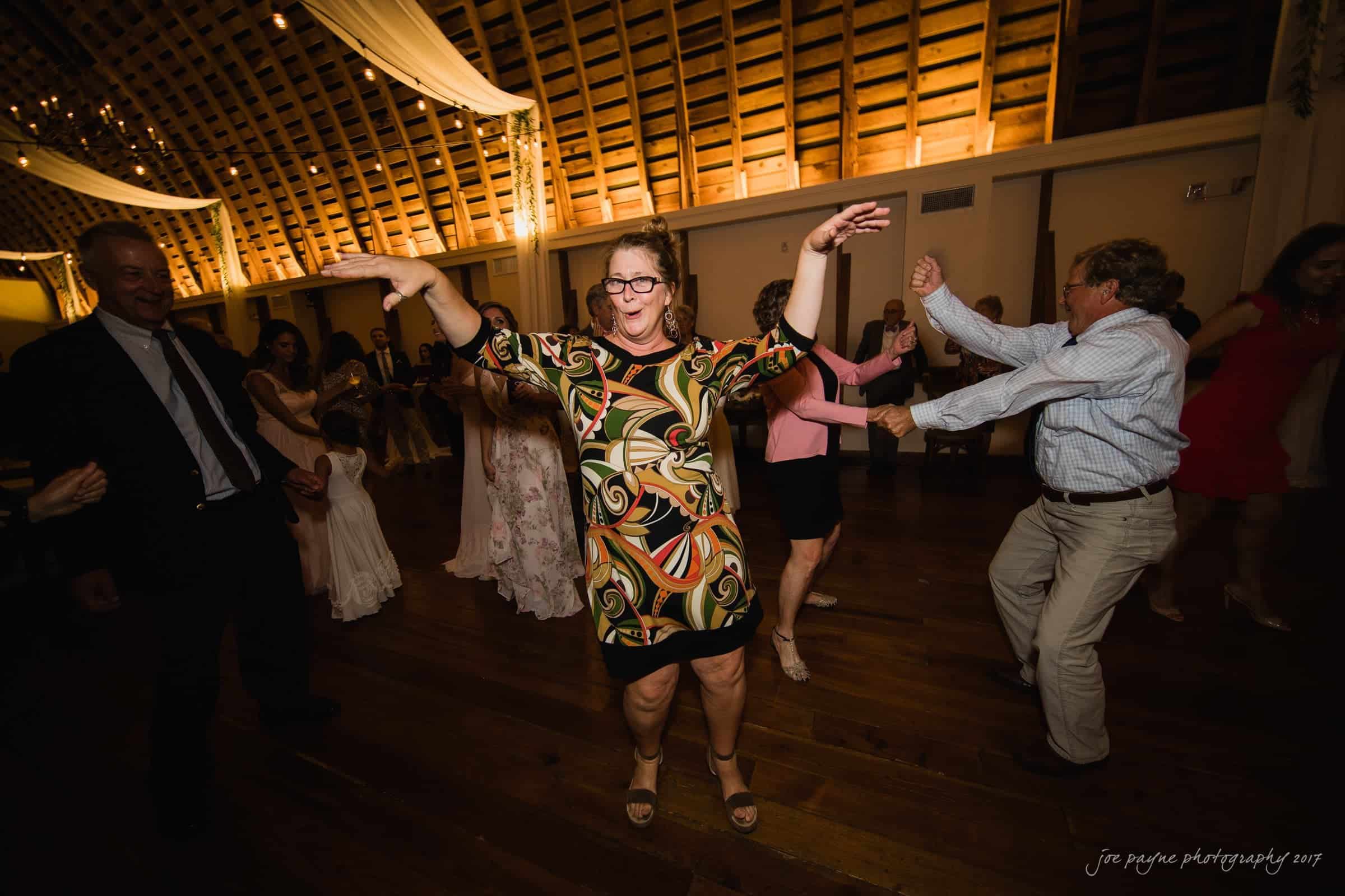 winmock nc wedding photographer - hallie & cullen A joyful dance floor scene at an event, with a woman in a colorful dress energetically dancing at the center, surrounded by other people enjoying themselves. The setting has a rustic, warmly lit, wooden interior.