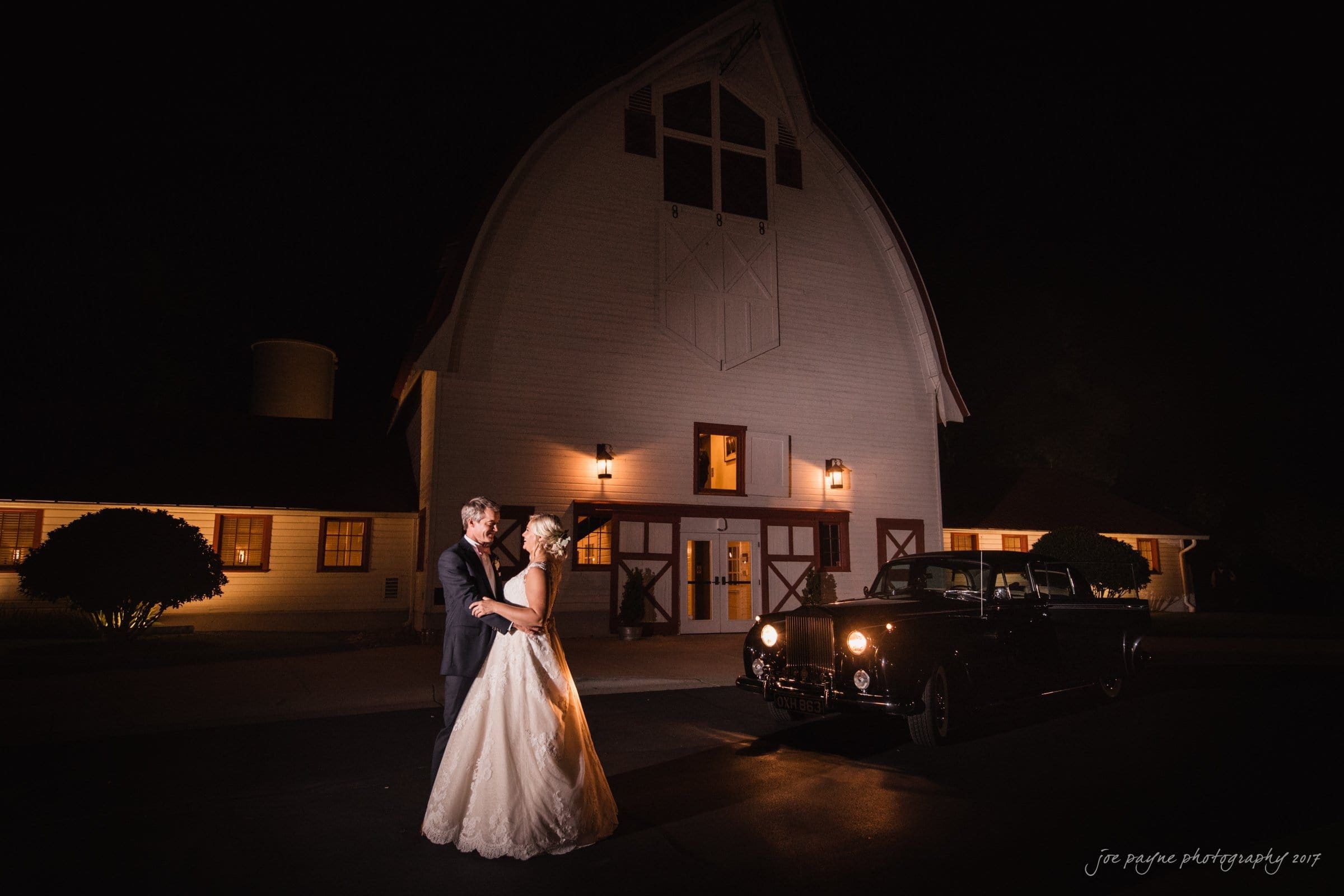 winmock nc wedding photographer - hallie & cullen A couple embraces on a dark night in front of a large illuminated barn. They are standing next to a vintage car with headlights on, adding a romantic glow to the scene.