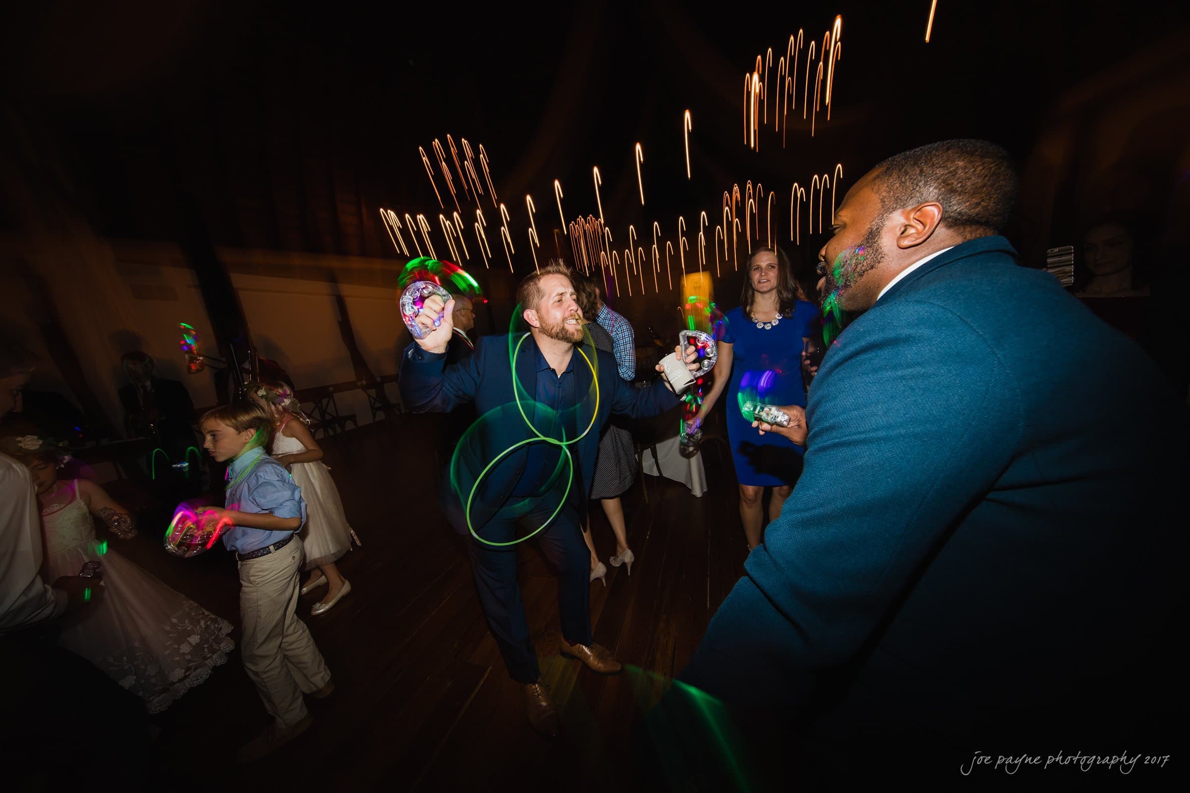 winmock nc wedding photographer - hallie & cullen A lively dance floor scene at a wedding, with guests joyfully dancing and holding glow sticks. A man in a suit energetically moves in the center while colorful lights and beams create a festive atmosphere.
