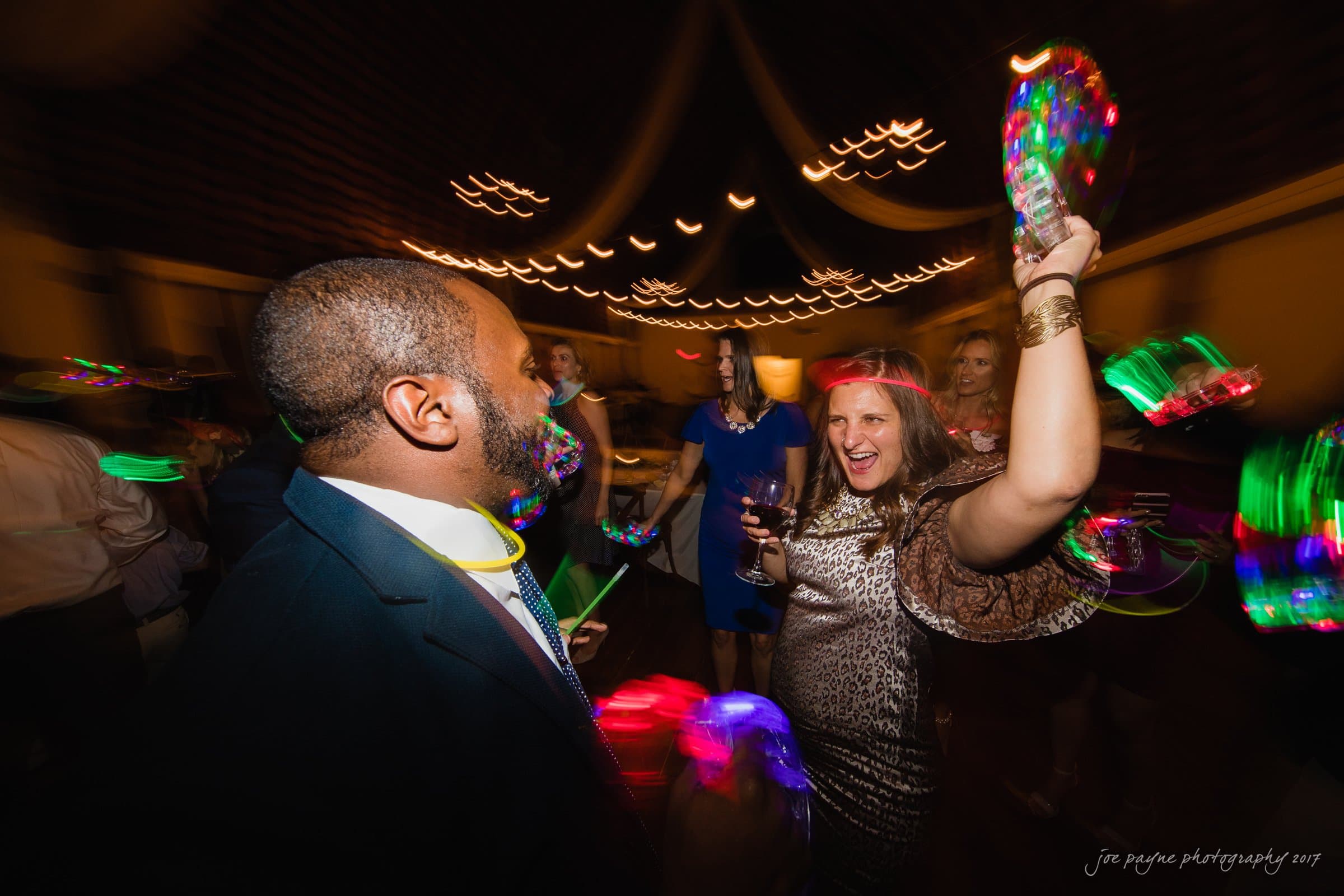 winmock nc wedding photographer - hallie & cullen A joyous group of people dancing at a party, with vibrant colorful lights and blurred motion capturing their energy. A woman in a leopard print dress enthusiastically raises her hand, while others enjoy the lively atmosphere.