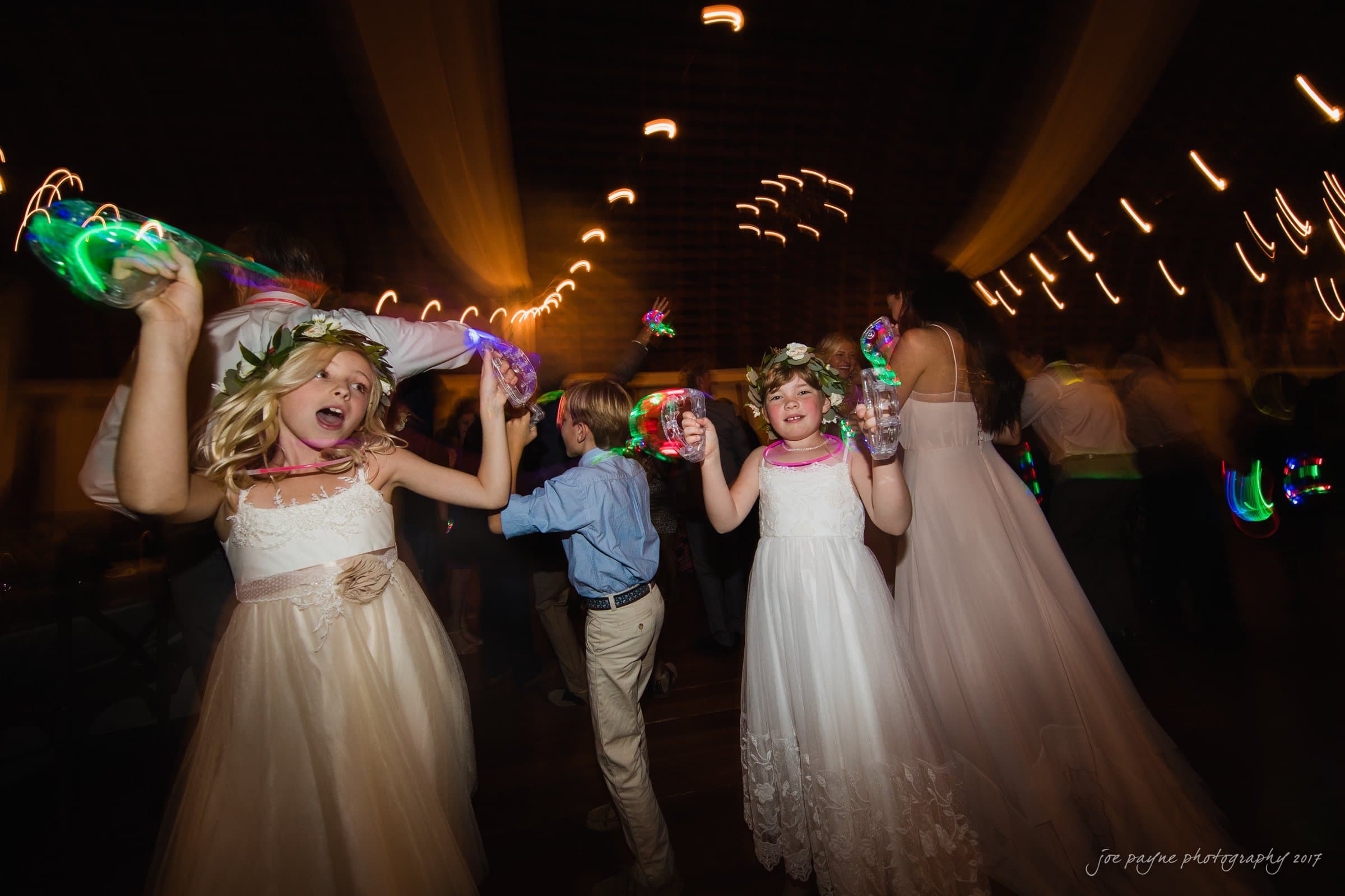 winmock nc wedding photographer - hallie & cullen Two young girls in white dresses dance energetically with colorful glow sticks at a lively event. A boy in a blue shirt joins the fun. Blur effects enhance the festive atmosphere with strings of lights in the background.