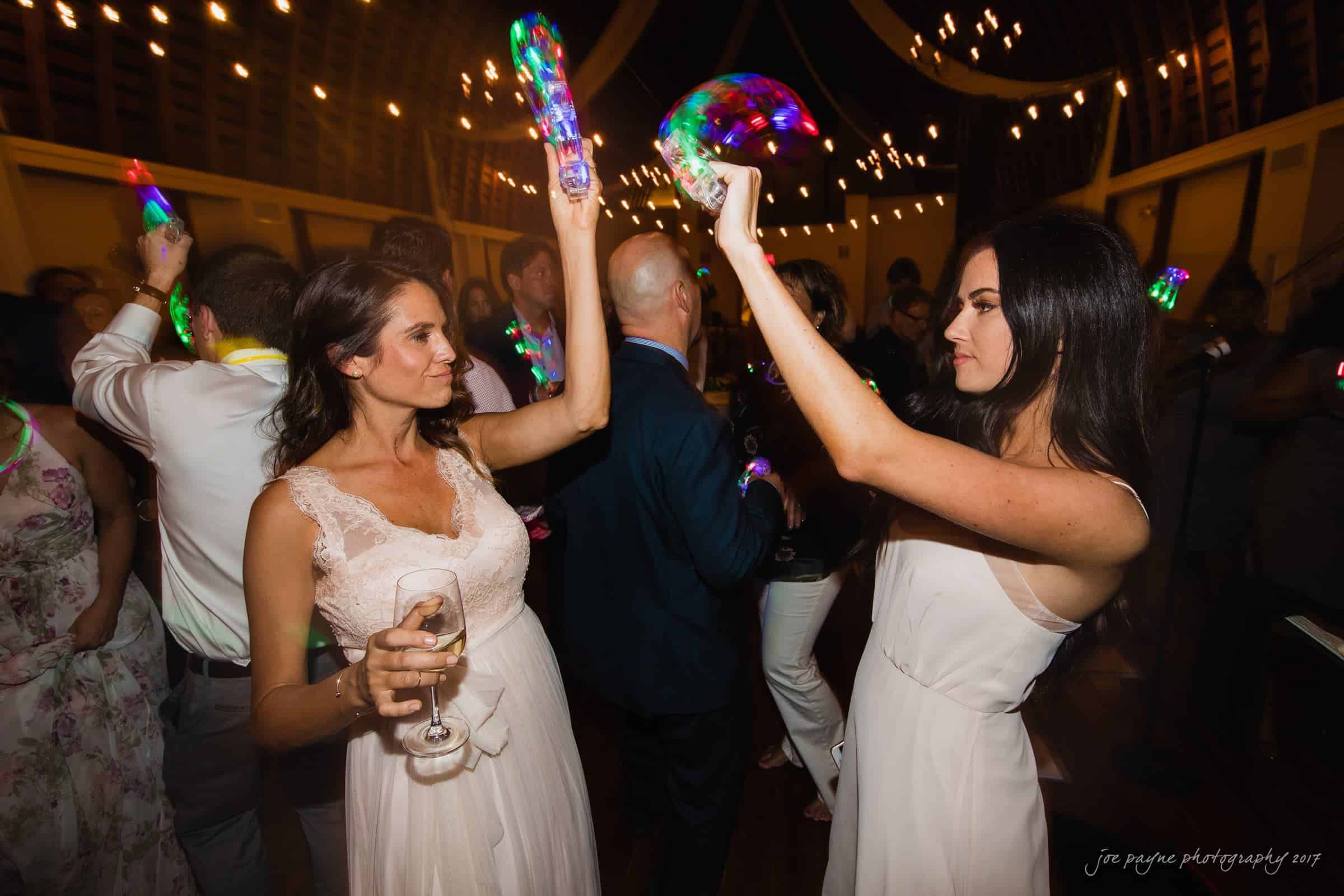 winmock nc wedding photographer - hallie & cullen Two women in dresses dance at a lively party. They are surrounded by colorful lights and other guests in a dimly lit room. One woman holds a glass, and the other waves her arms in the air, capturing a festive atmosphere.