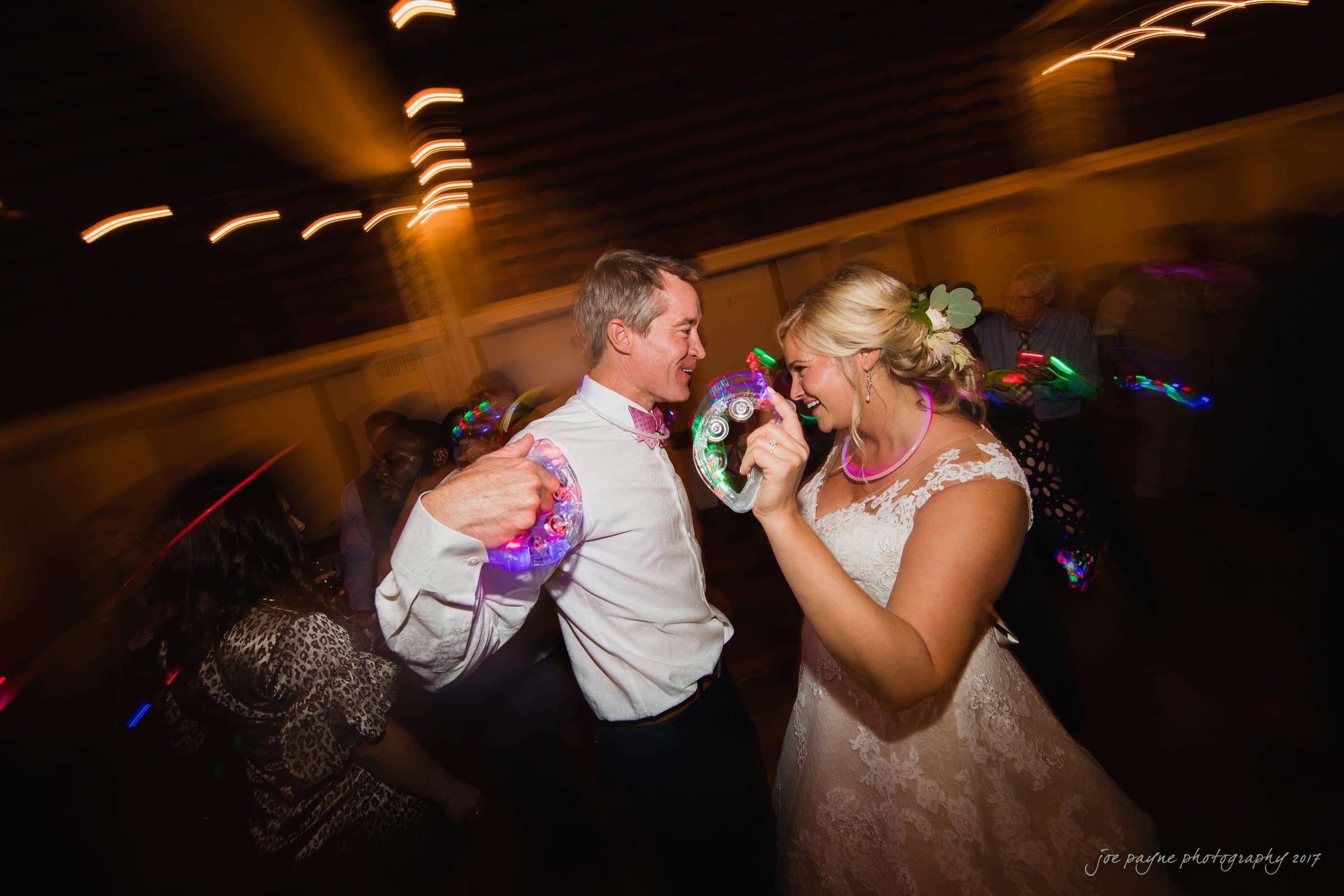 winmock nc wedding photographer - hallie & cullen A bride and groom dance energetically at a wedding reception, surrounded by colorful lights. The bride wears a lace dress and flower in her hair, while the groom wears a shirt and bow tie. They hold glowing tambourines, creating a lively atmosphere.
