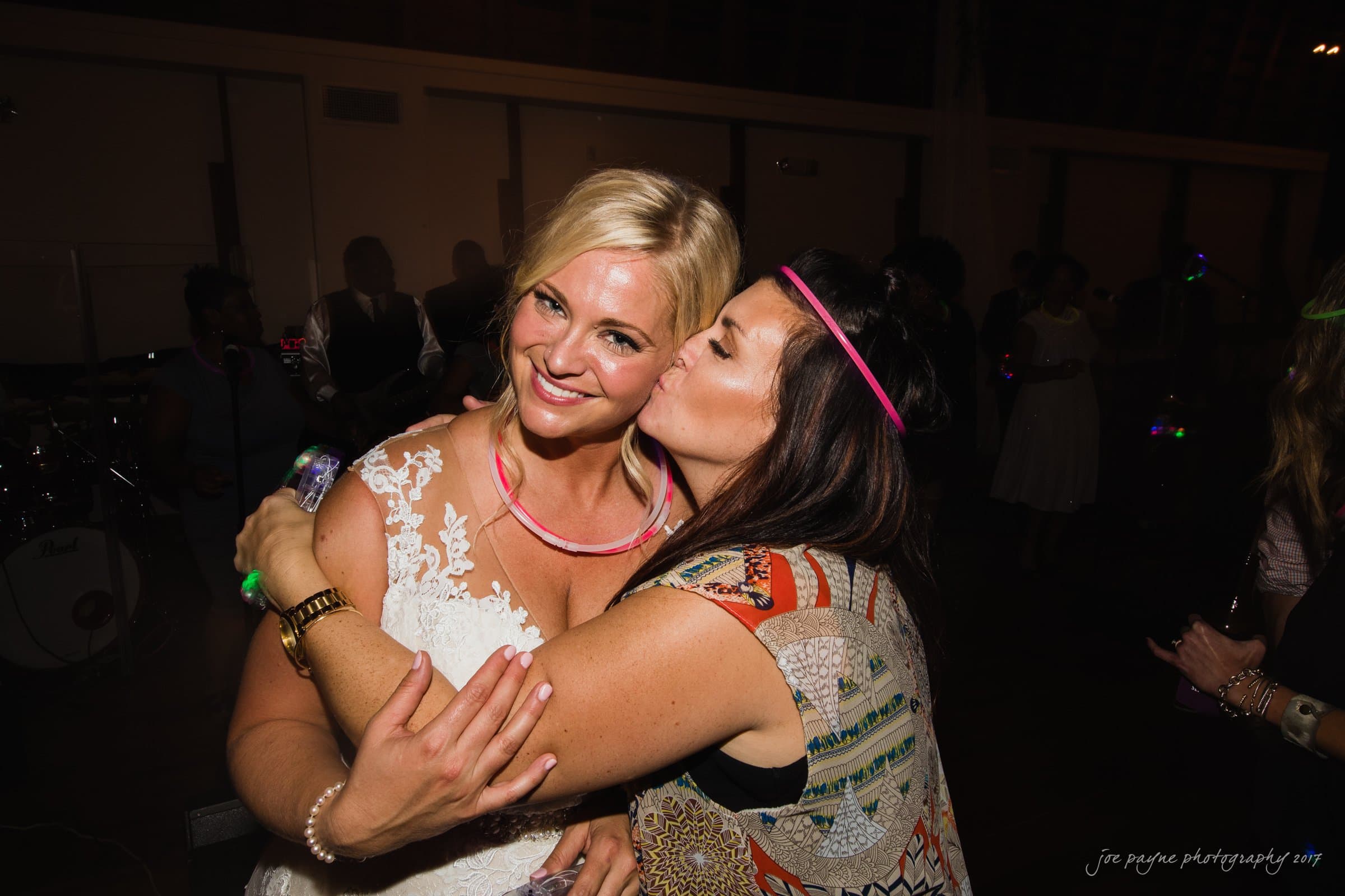 winmock nc wedding photographer - hallie & cullen A bride with blonde hair smiles as she is hugged by another woman who is kissing her cheek. Both are wearing glow necklaces in a dimly lit room, suggesting a joyful celebration or wedding reception.
